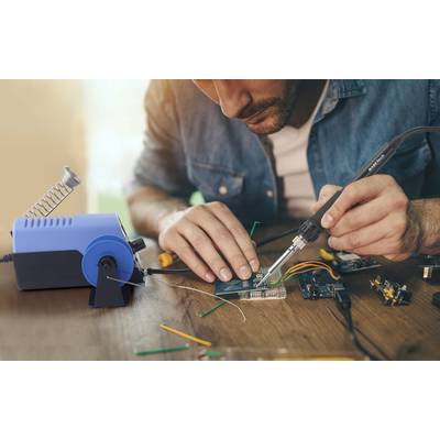 Mann lötet konzentriert eine Leiterplatte auf einem Tisch, mit Werkzeugen und Lötgerät daneben. Elektronik und Schaltkreise sichtbar.