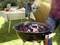 Eine Person entzündet Kohle in einem Grill mit einem Heißluftgebläse. Im Hintergrund steht ein Tisch mit Lebensmitteln im Garten.