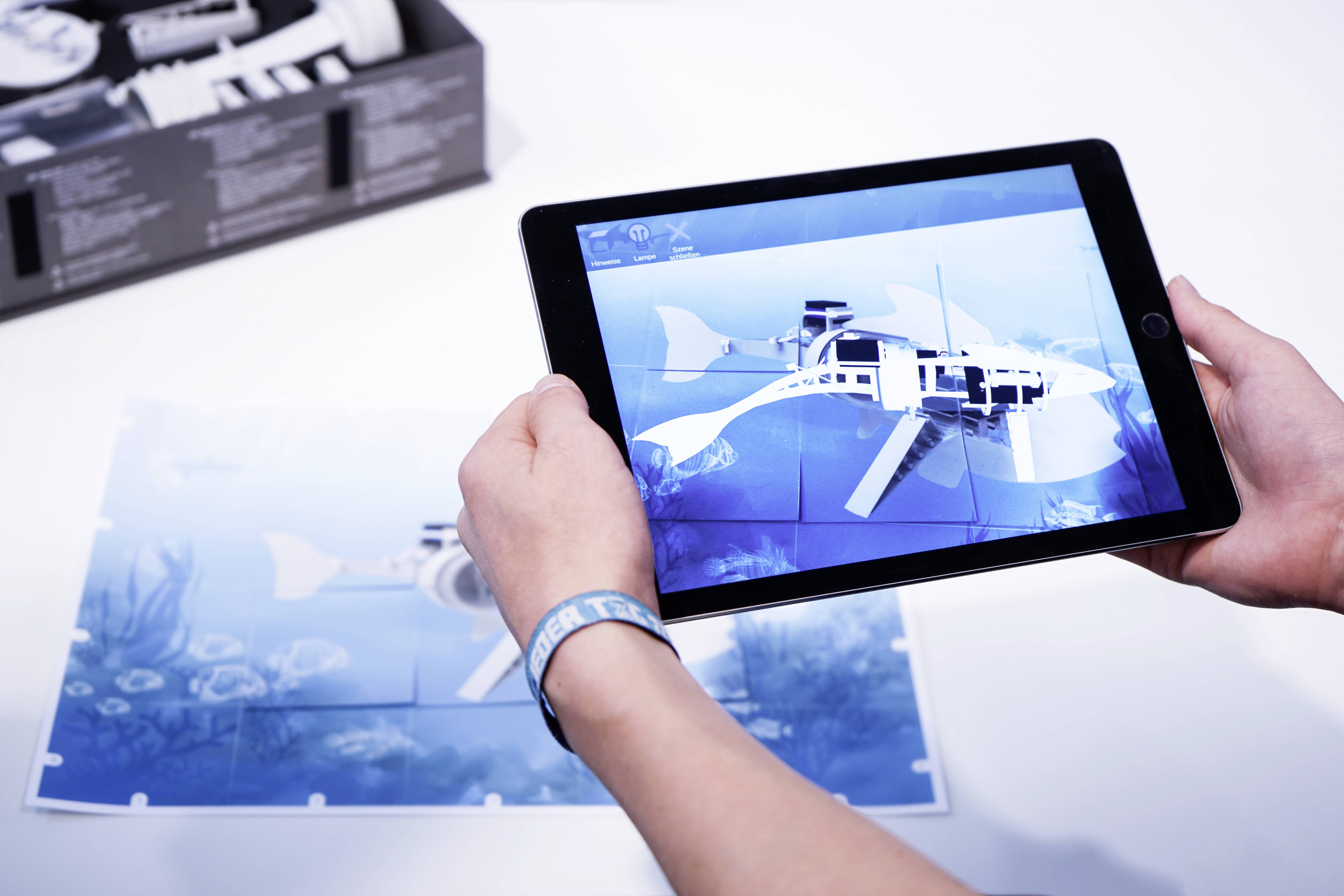 Eine Person hält ein Tablet, das ein digitales Modell eines Fisches zeigt, über Papierfragmenten. Im Hintergrund ist Kartonverpackung.