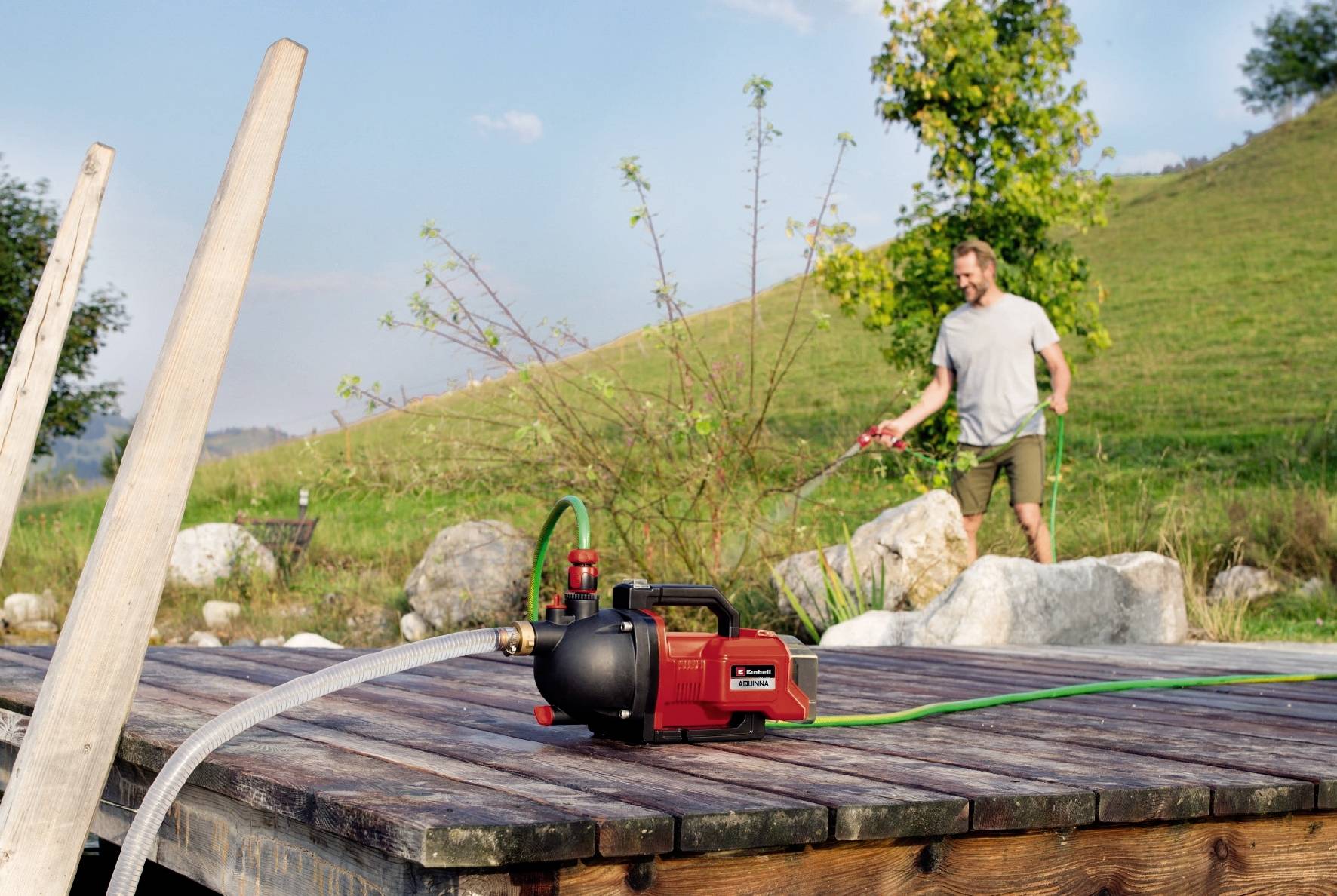 Ein Mann bewässert Pflanzen im Garten mit einem Gartenschlauch. Eine rote Wasserpumpe steht auf einer Holzfläche im Vordergrund.