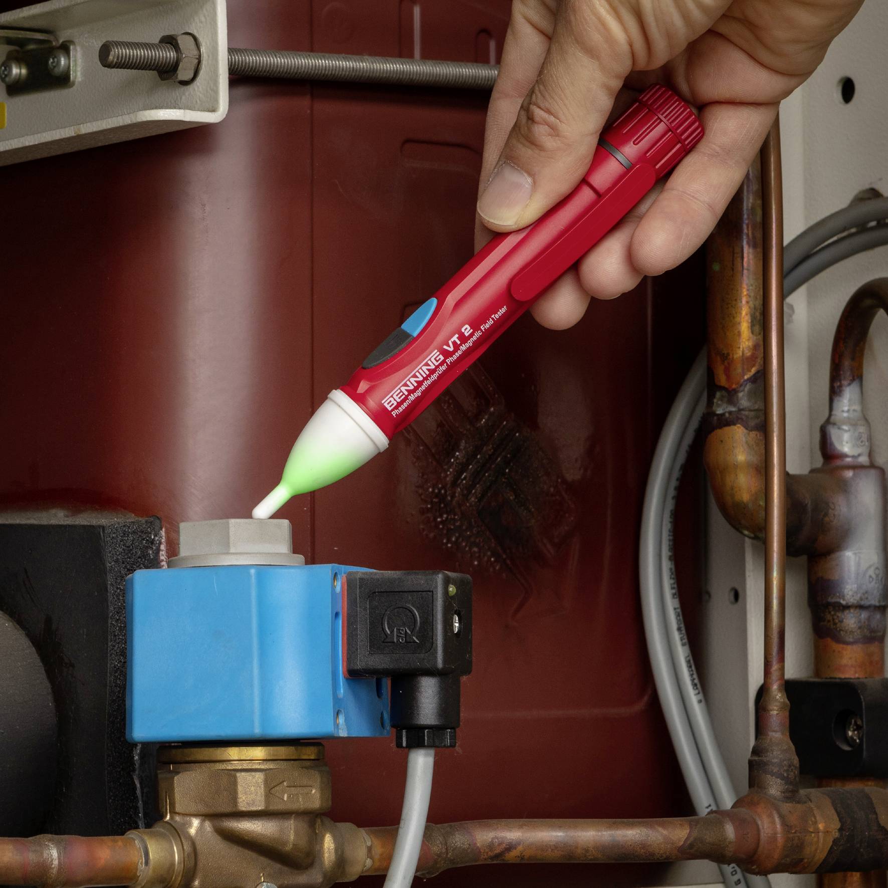 Eine Person hält ein rotes Testgerät über ein blaues Rohrventil, um Auslaufen oder elektrische Spannung zu prüfen.