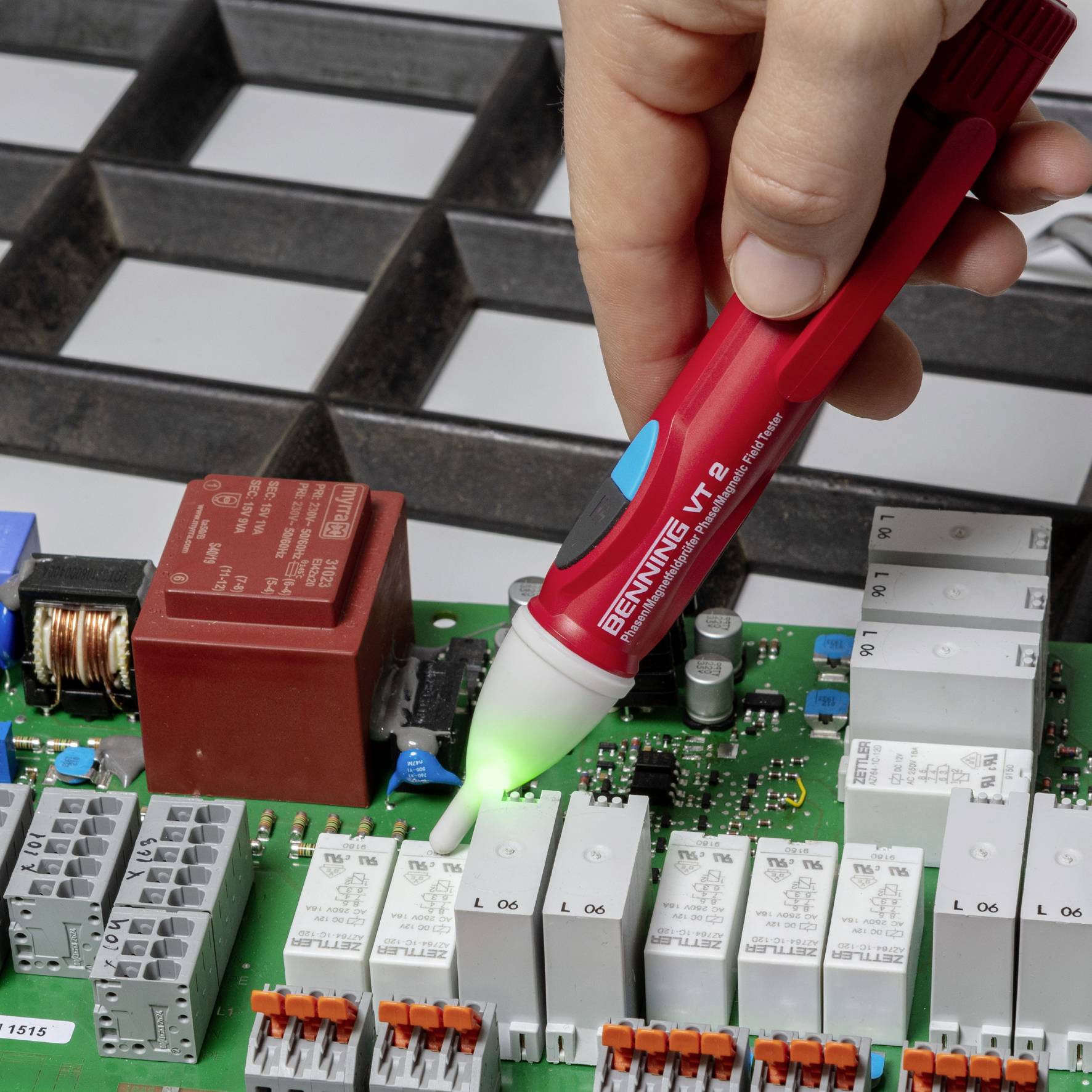 Eine Hand hält einen Spannungsprüfer über eine Leiterplatte, umgeben von elektrischen Bauteilen in einer technischen Umgebung.