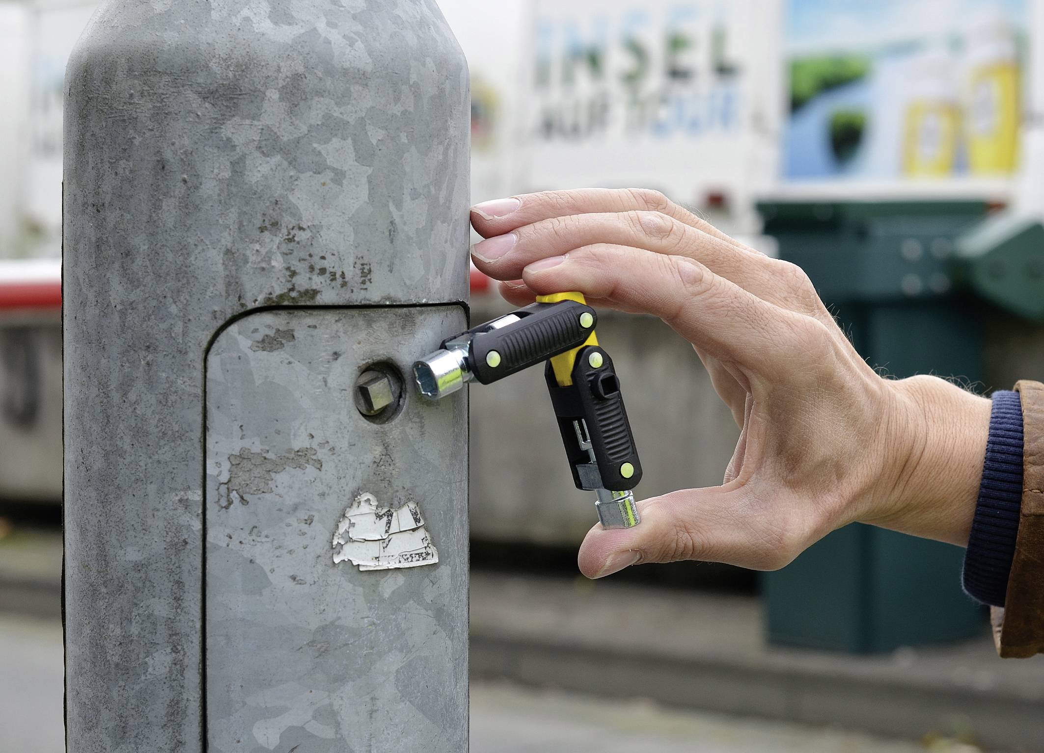 Eine Hand hält ein faltbares Fahrradschloss, das an einem Pfosten befestigt wird. Im Hintergrund ist unscharf eine Straße erkennbar.