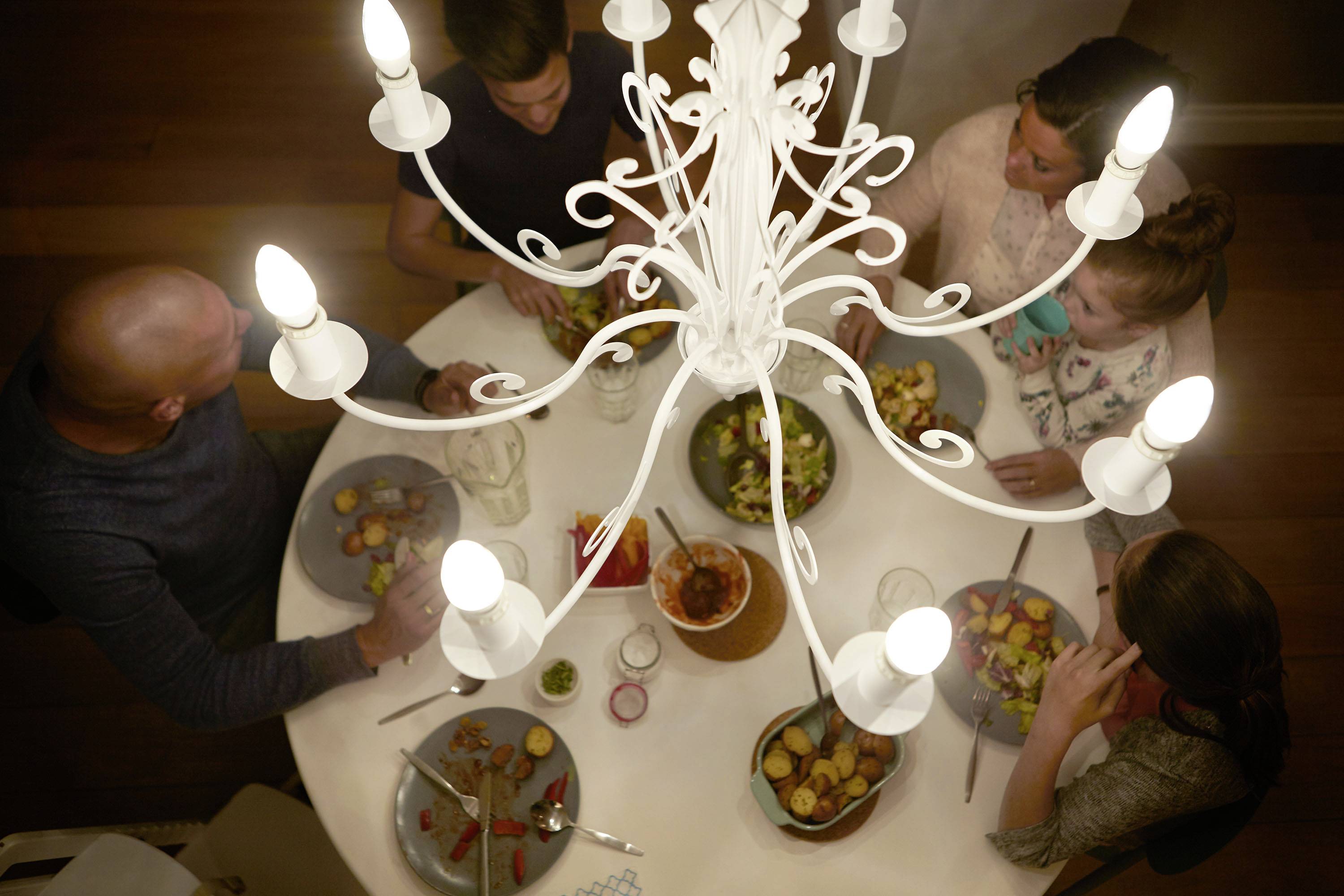 Fünf Personen essen gemeinsam Abendessen um einen runden Tisch unter einem Kronleuchter. Der Tisch ist mit verschiedenen Gerichten gedeckt.