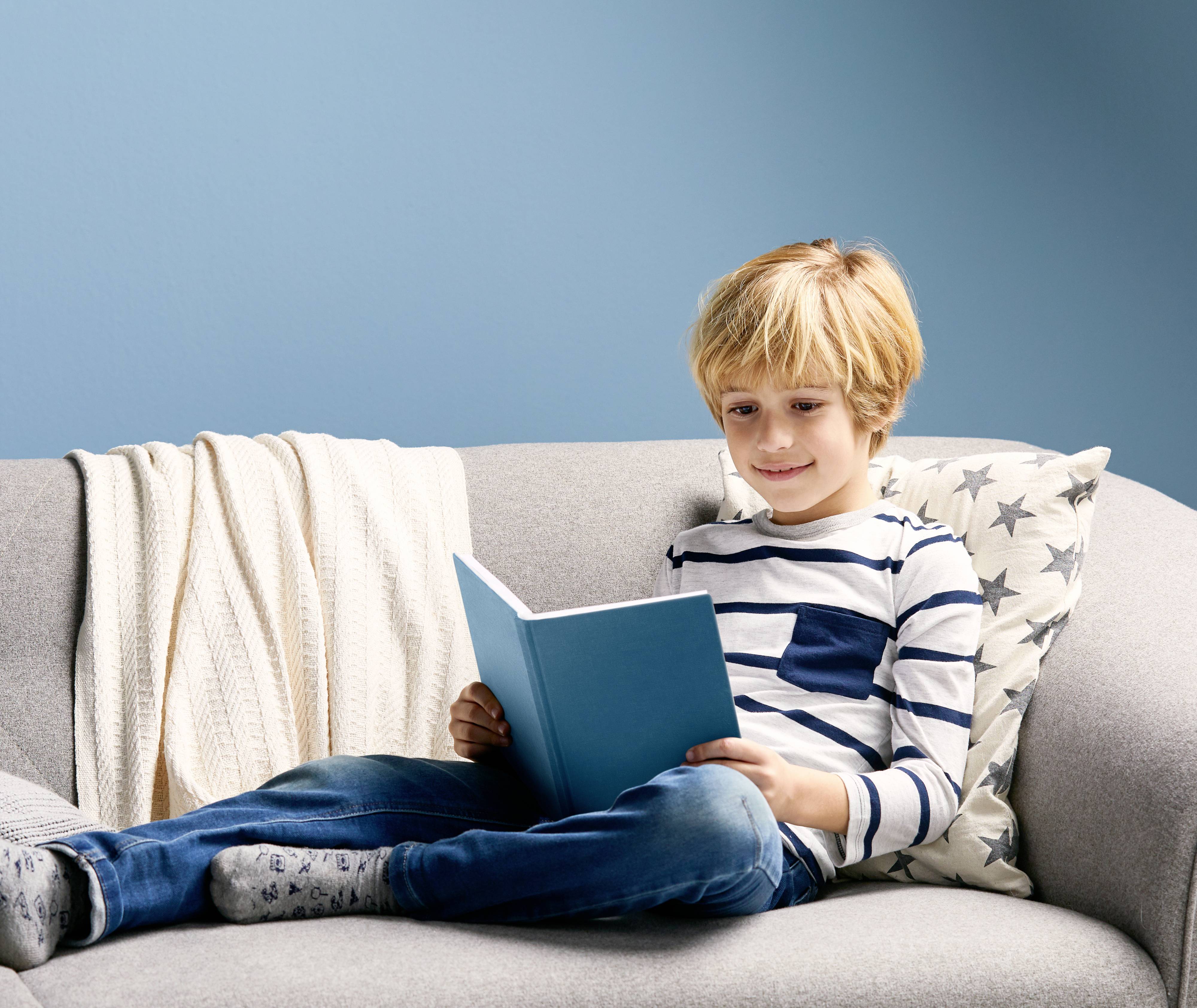 Ein Junge liest ein blaues Buch auf einem grauen Sofa. Er trägt ein gestreiftes Shirt und eine Jeans. Im Hintergrund ist eine blaue Wand.