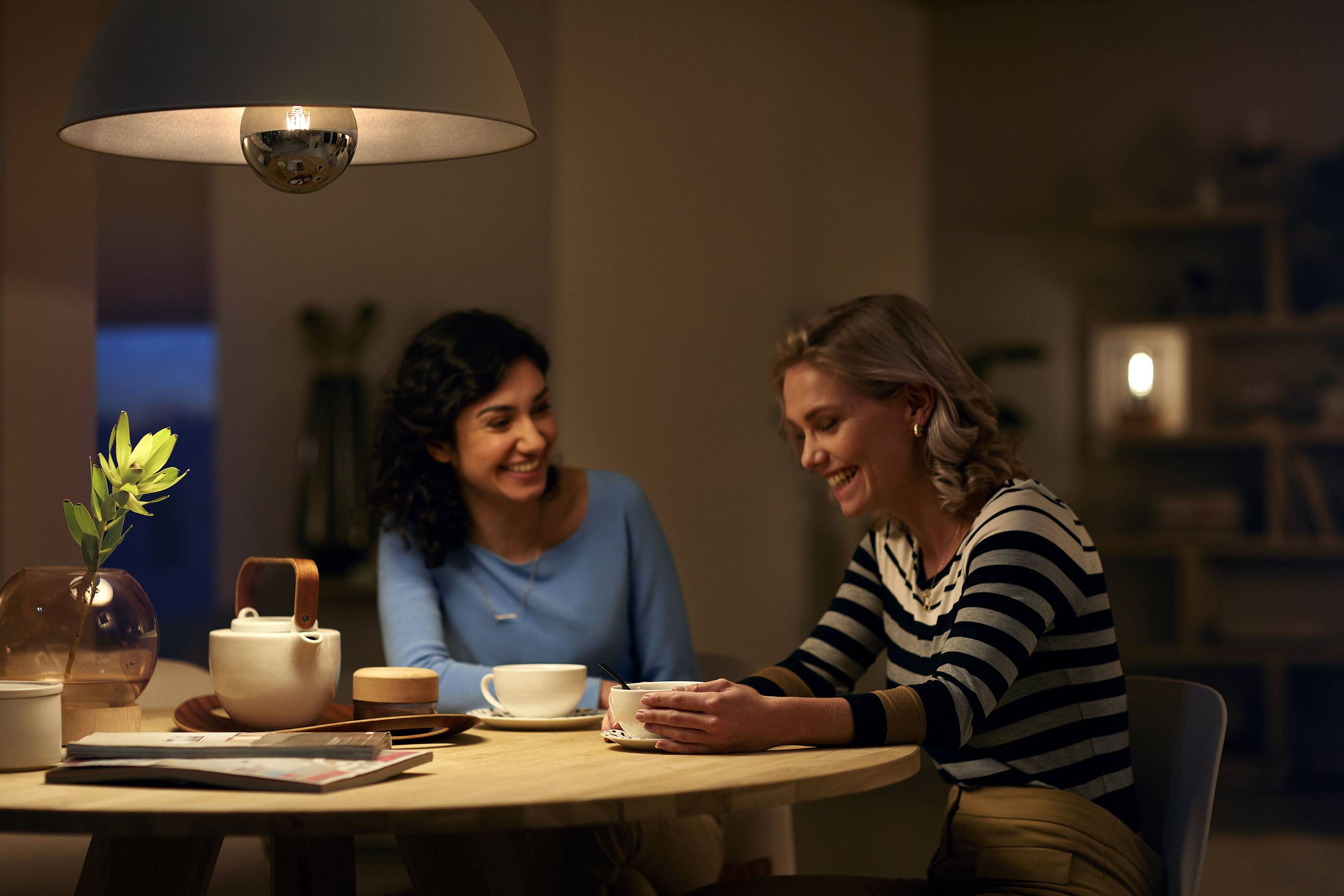 Zwei Frauen sitzen lächelnd an einem Tisch mit Teetassen und einem Teeservice, in einem gemütlich beleuchteten Raum.