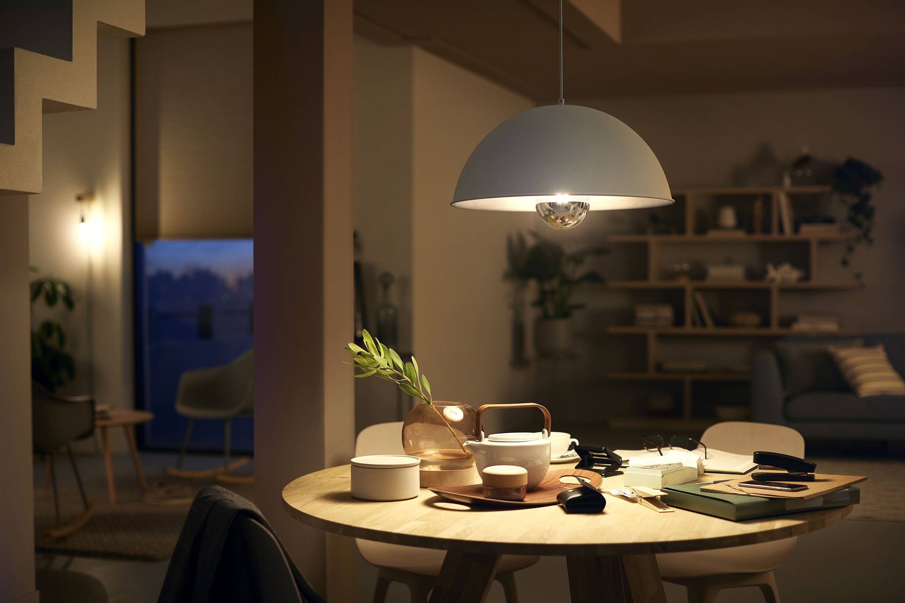 Wohnzimmer mit einem runden Esstisch, auf dem Geschirr, Bücher und Pflanzen stehen. Im Hintergrund ein Sofa und Regal. Gedämpfte Beleuchtung.