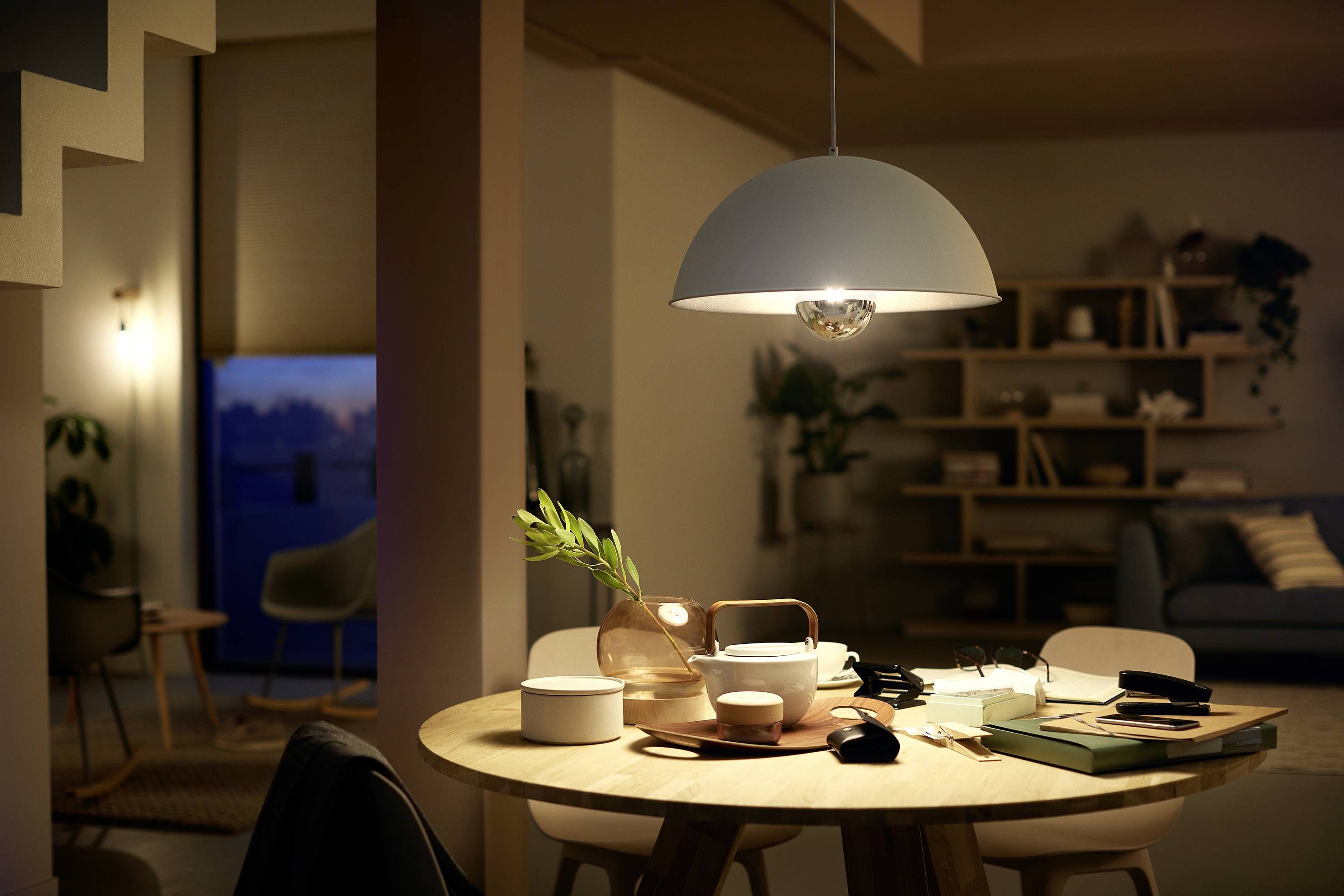 Ein Tisch mit Geschirr und Büchern, beleuchtet von einer Hängelampe. Im Hintergrund ein Wohnzimmer mit Regalen und Sofas.