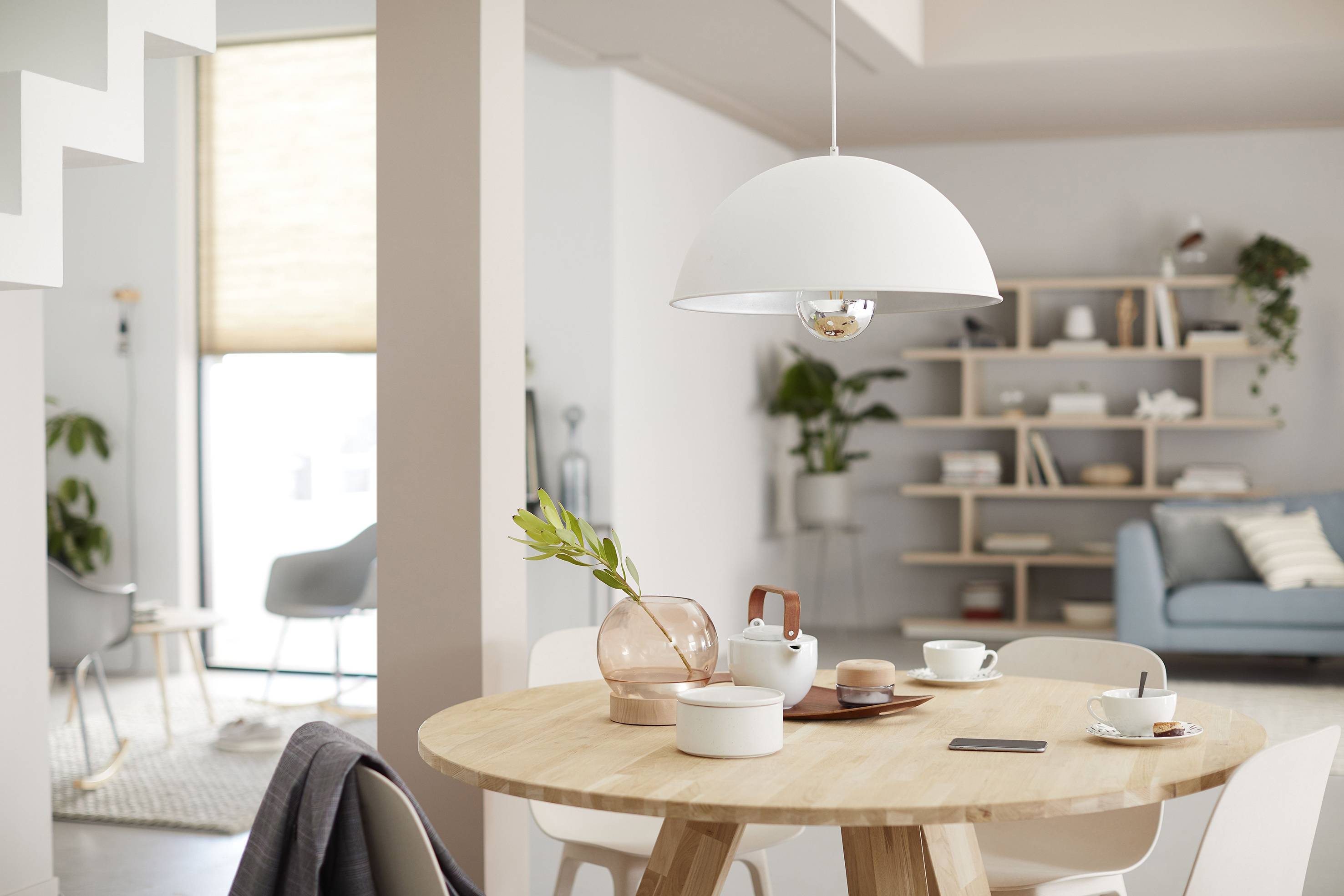 Ein helles Wohnzimmer mit Esstisch im Vordergrund, auf dem Geschirr und eine Vase mit grünen Zweigen stehen. Im Hintergrund ein Sofa und ein Regal.