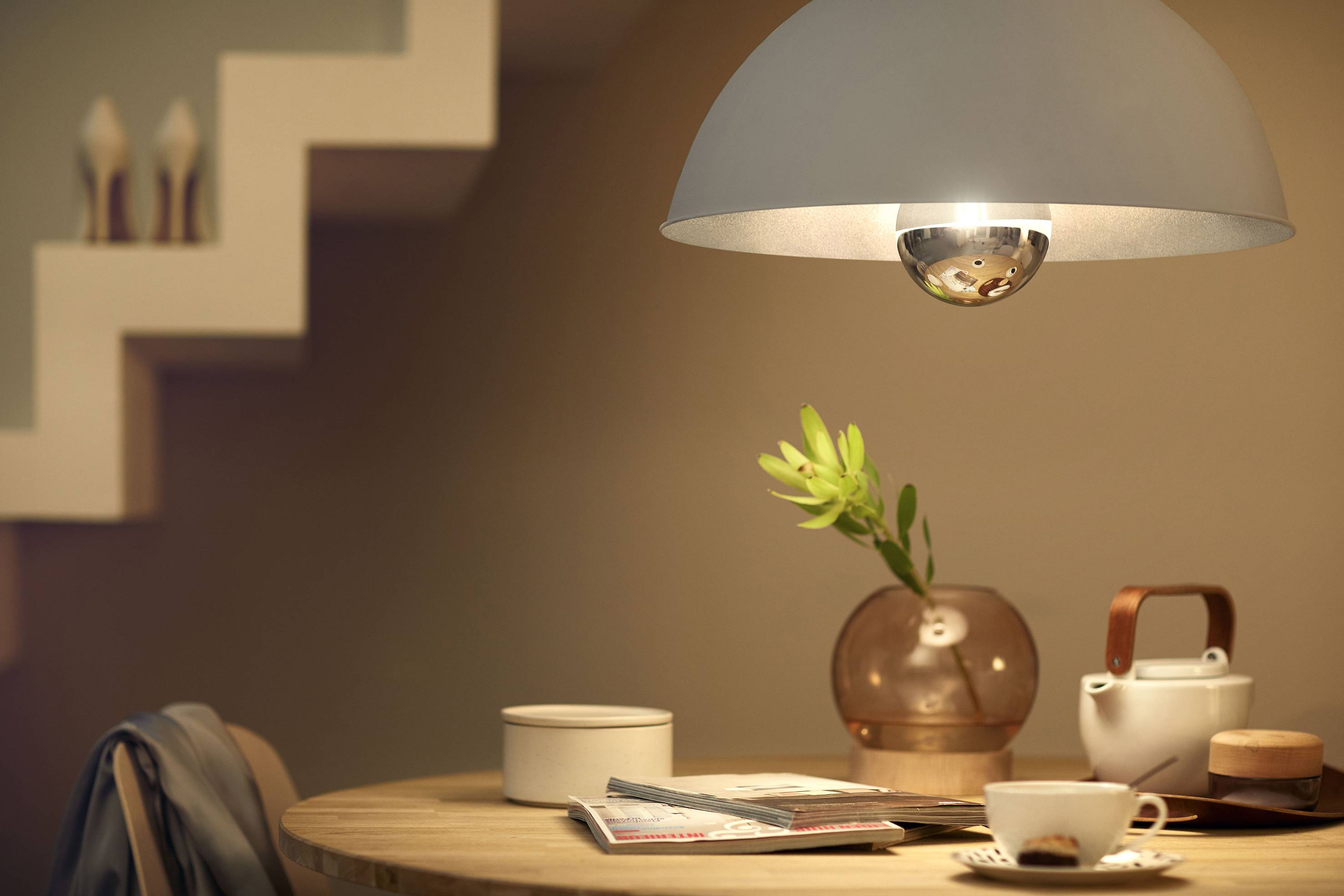 Gemütliche Tischszene mit Teekanne, Tasse, Vase mit Pflanzenzweig und Zeitschrift unter einer modernen Hängelampe.