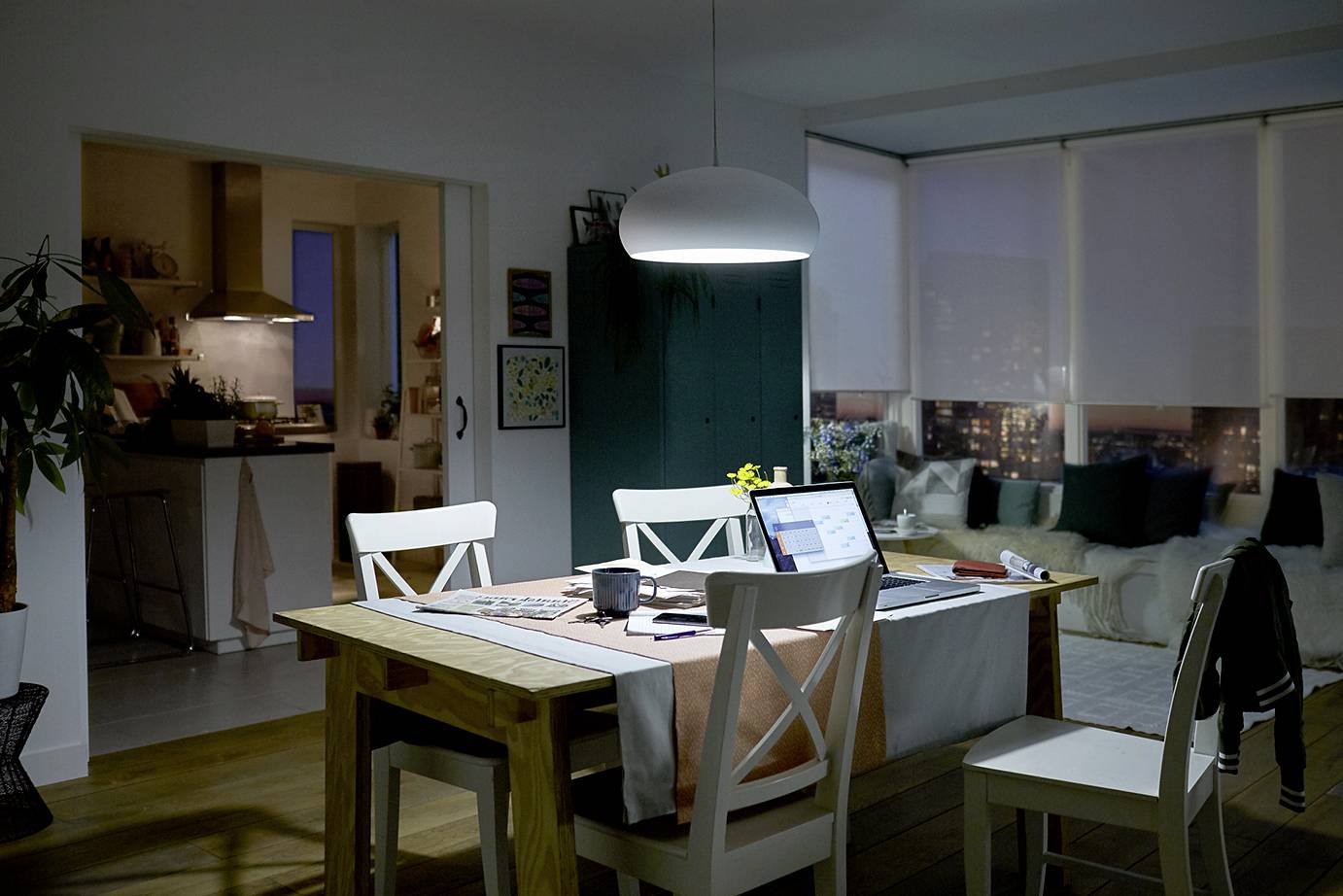 Ein Wohnzimmer bei Nacht mit einem Tisch im Vordergrund. Auf dem Tisch sind ein Laptop, ein Notizbuch und eine Kaffeetasse. Im Hintergrund eine Küche.