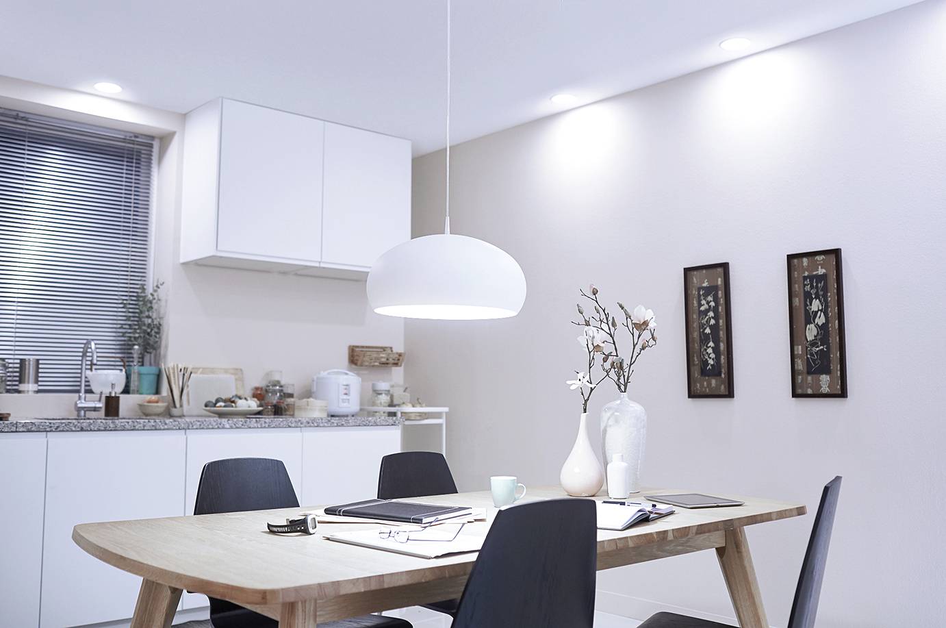 Wohnzimmer mit Esstisch, umgeben von vier schwarzen Stühlen. Weiße Küchenschränke und Dekorationen im Hintergrund, helle Wandbeleuchtung.