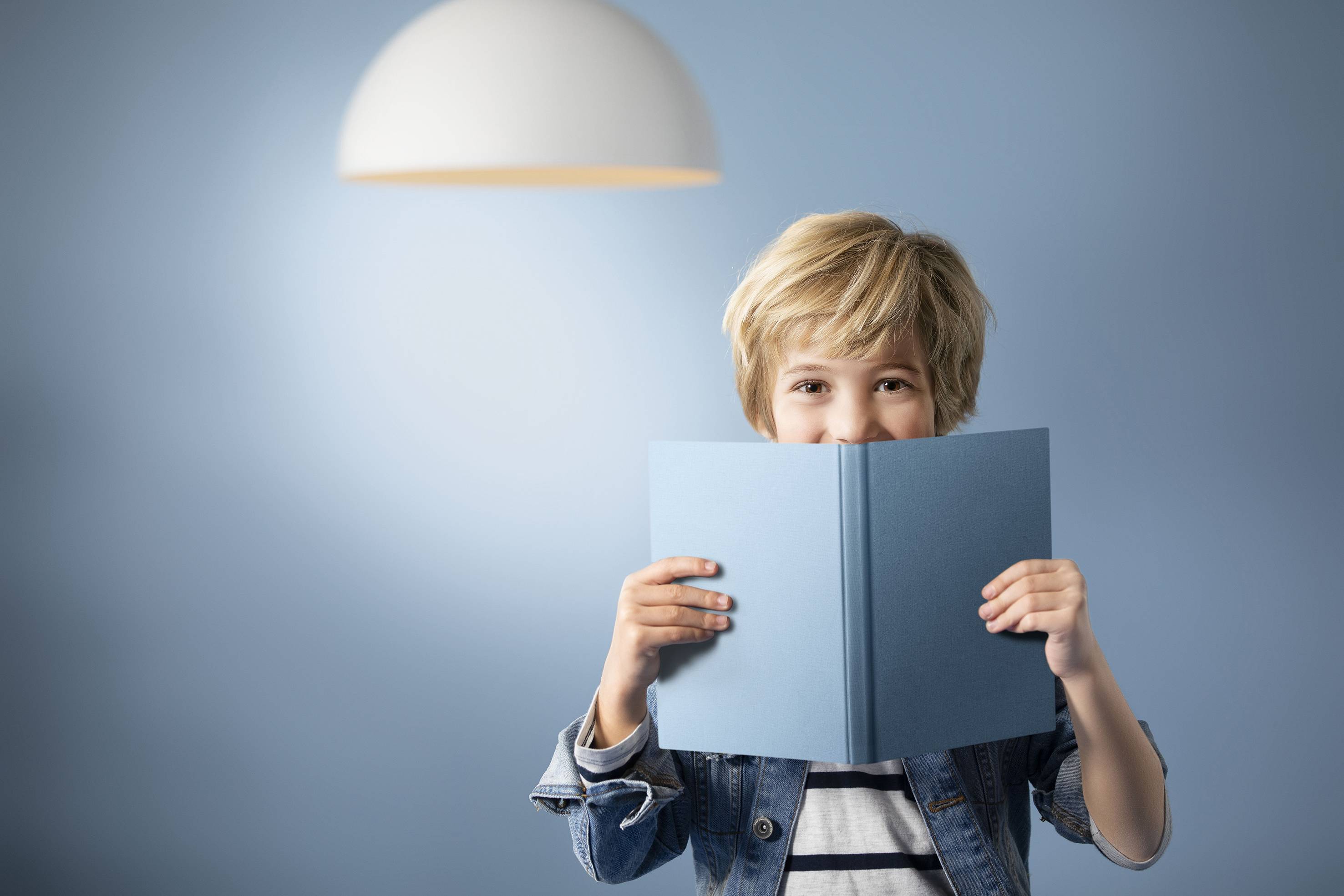 Ein Kind mit blonden Haaren hält ein geöffnetes blaues Buch vor sich. Im Hintergrund ist eine blaue Wand mit einer hängenden Lampe.