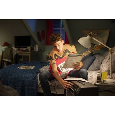 Ein Teenager sitzt auf einem Bett, hält eine E-Gitarre und liest Notenblätter. Eine Lampe leuchtet auf ihn und ein Verstärker steht in der Nähe.