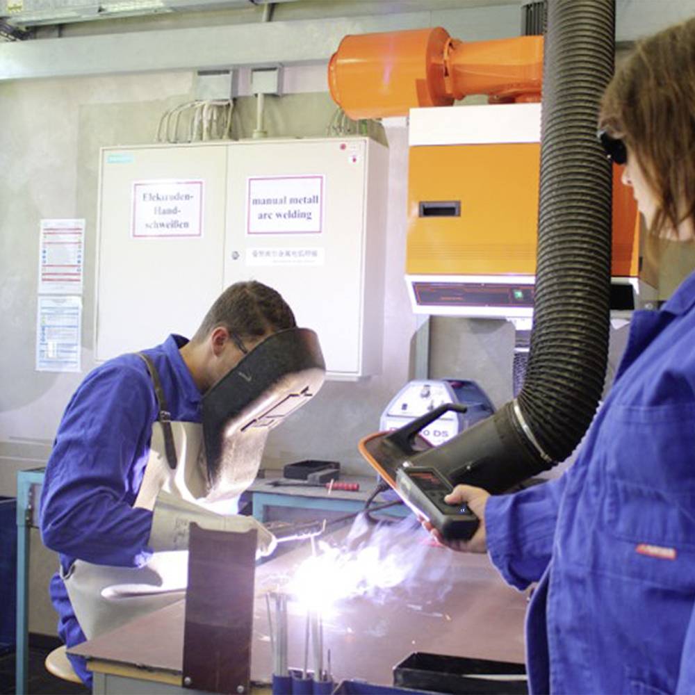 Ein Mann in blauer Arbeitskleidung schweißt Metallstücke zusammen, während ein anderer Arbeiter eine Schweißmaschine überwacht.