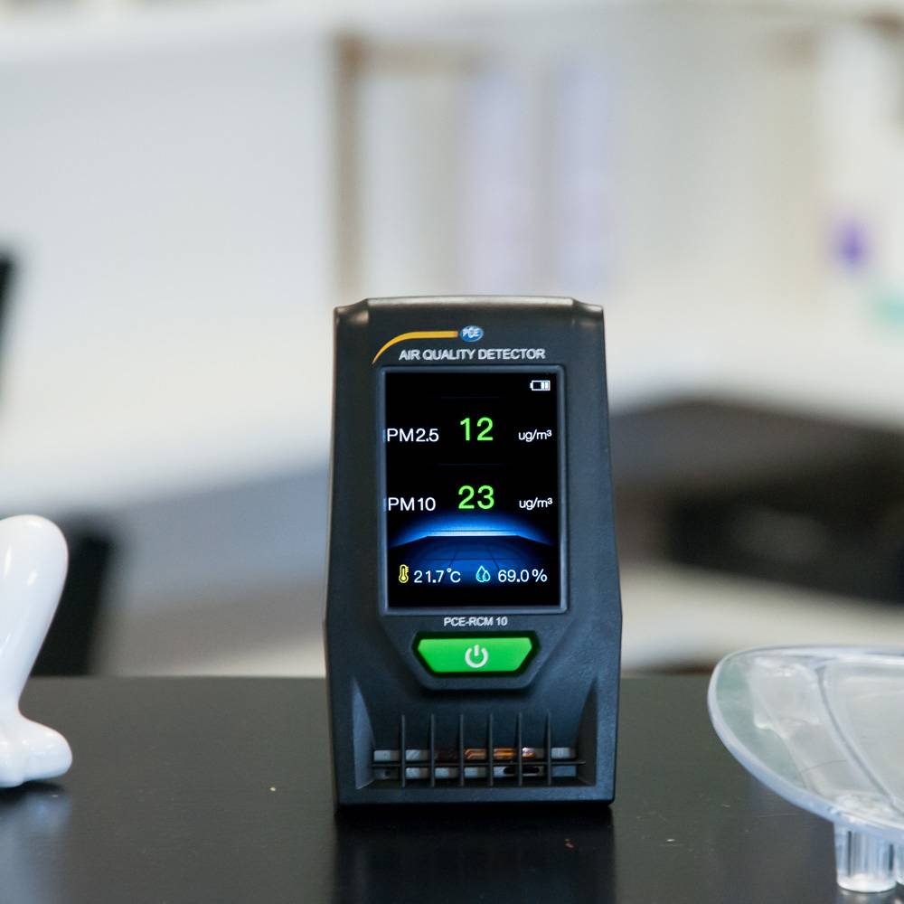 Ein Luftqualitätsdetektor zeigt PM2.5 bei 16 µg/m³ und PM10 bei 23 µg/m³ an, was gute Luft anzeigt. Gerät steht auf einem Tisch.