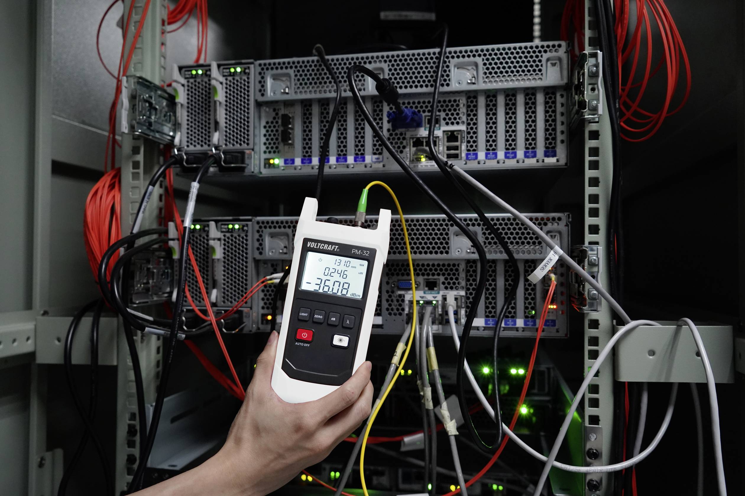 Ein Hand hält ein Messgerät, das die Temperatur in einem Serverraum anzeigt. Im Hintergrund sind mehrere Server mit Kabeln zu sehen.