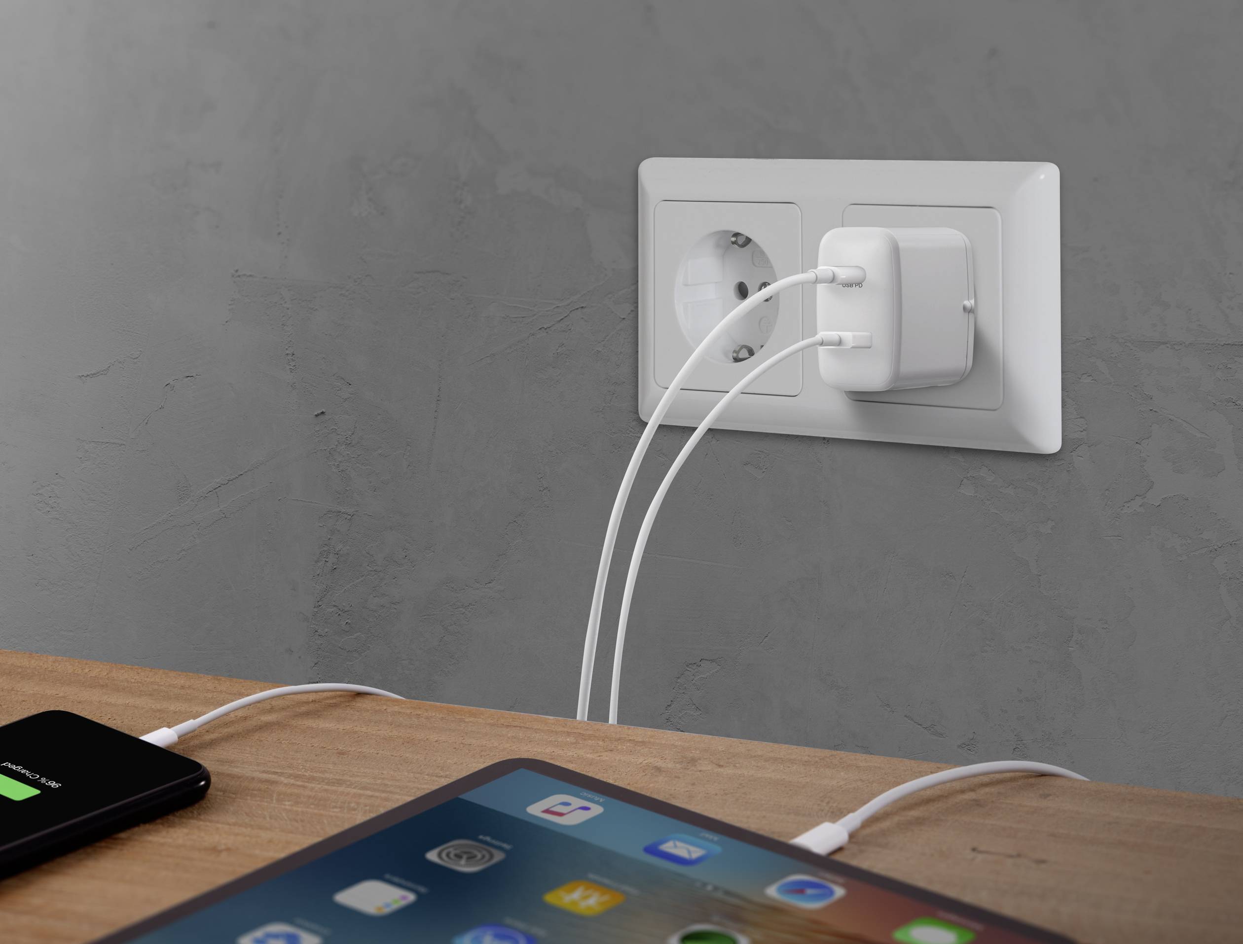 Ein Tisch mit einem Smartphone und einem Tablet, die an einer Steckdose an der Wand aufgeladen werden. Beide Geräte sind eingeschaltet.