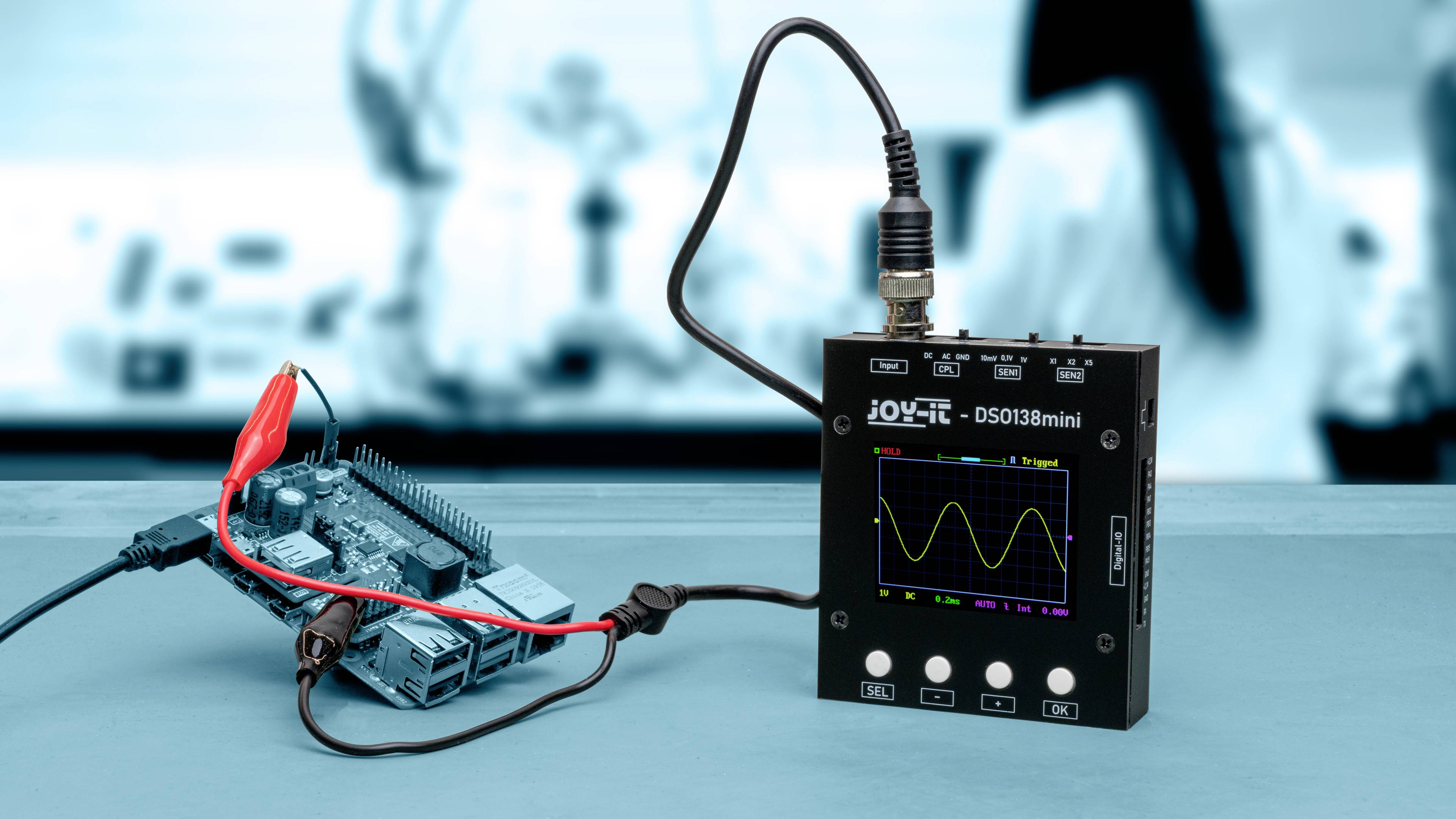 'Oszilloskop DS0138mini' zeigt Sinuskurve auf Display, verbunden mit Mikrocontrollerplatine über rote und schwarze Kabel. Im Laborhintergrund.