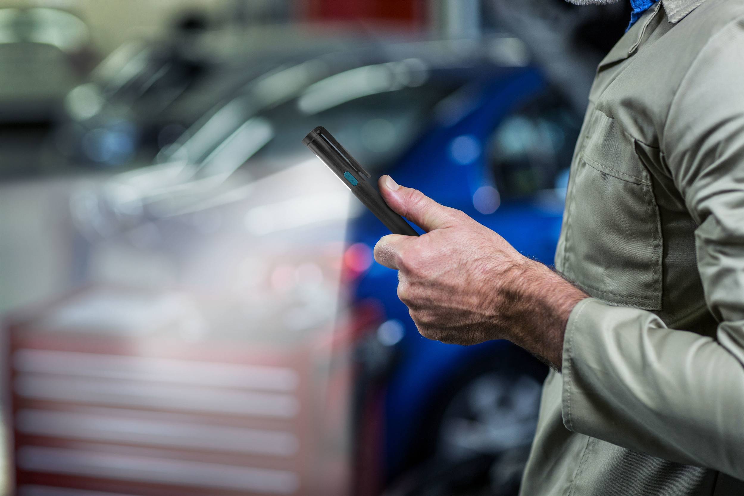 Eine Person in Arbeitskleidung hält ein Smartphone in einer Autowerkstatt. Im Hintergrund ist unscharf ein blaues Auto zu sehen.