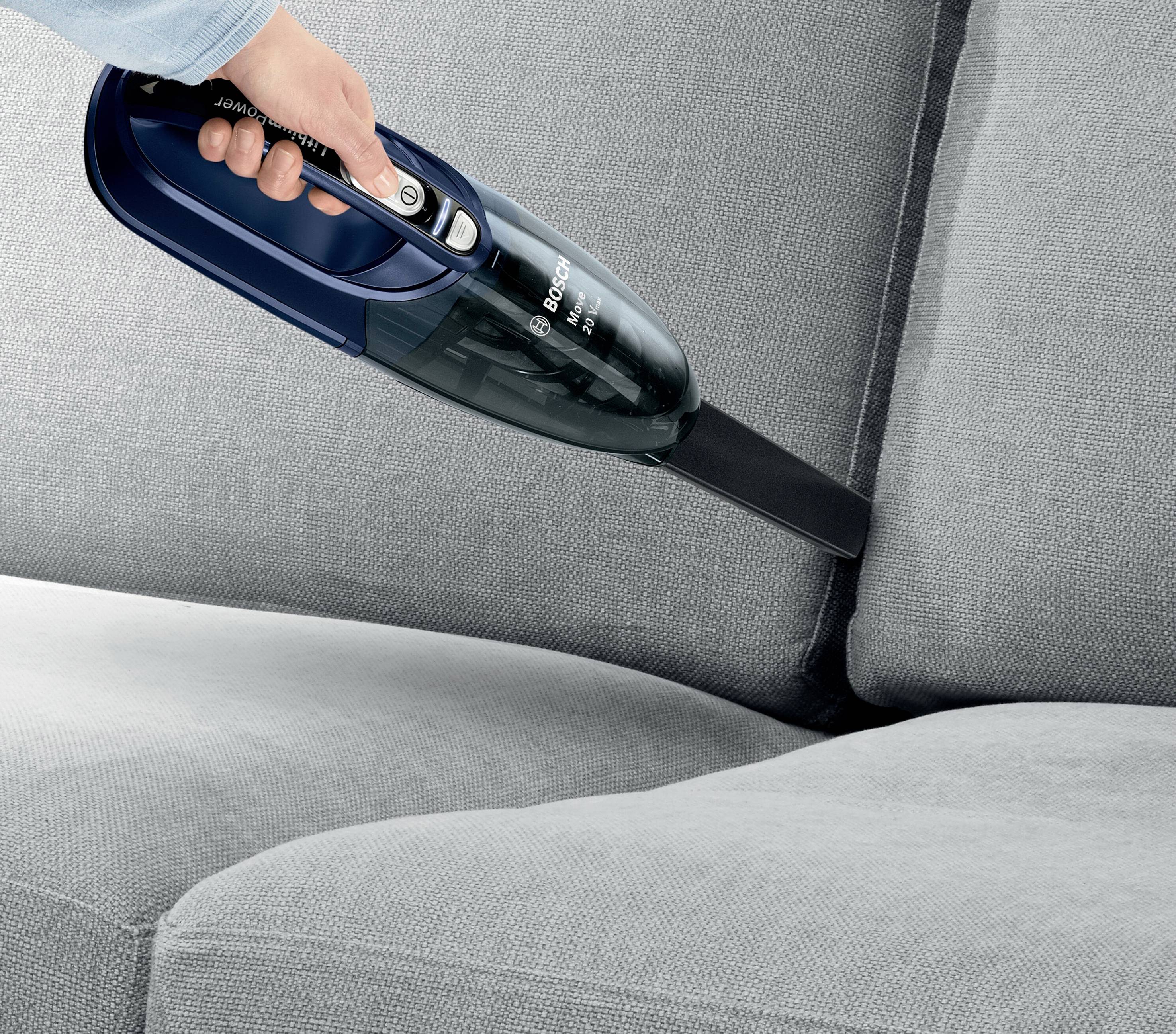 Eine Person reinigt mit einem Handstaubsauger die Spalte eines grauen Sofas.