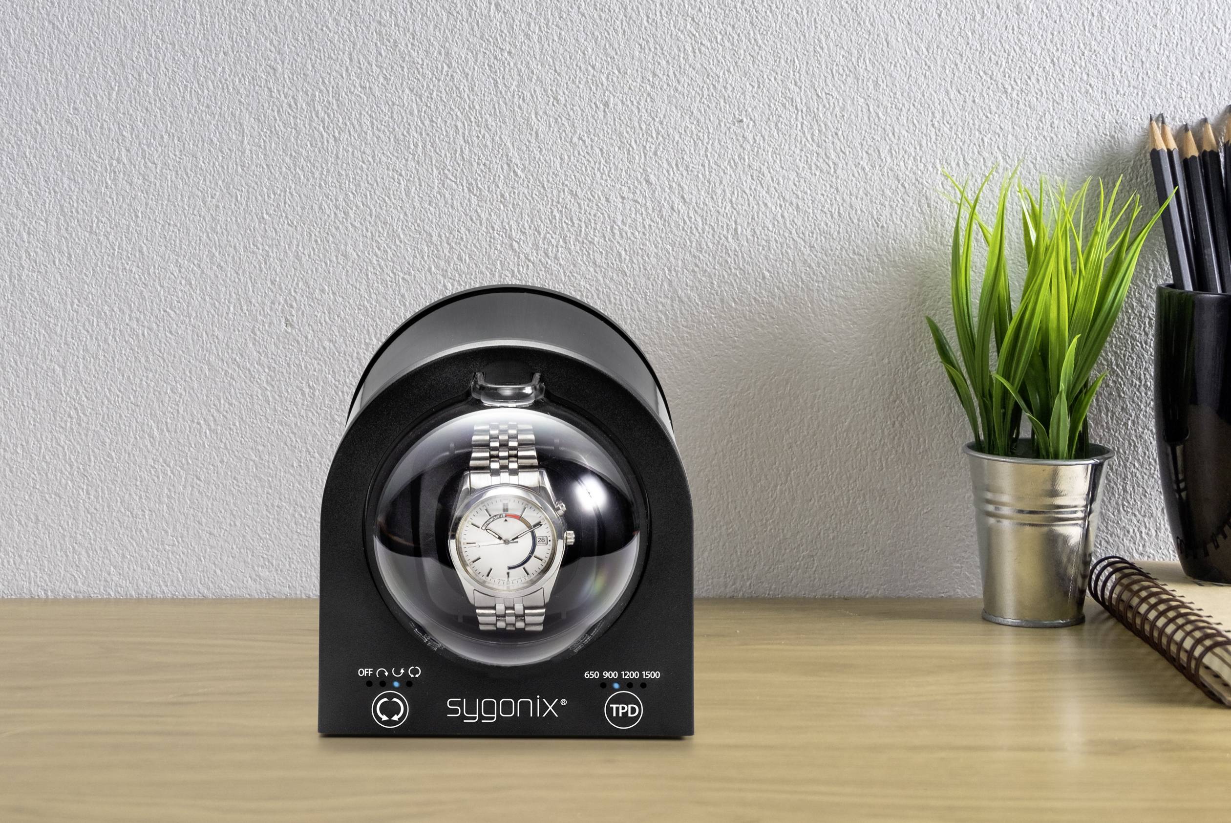 Uhrenbeweger auf einem Tisch mit silberner Uhr im Inneren, daneben eine Pflanze und ein Notizbuch mit Stiften.