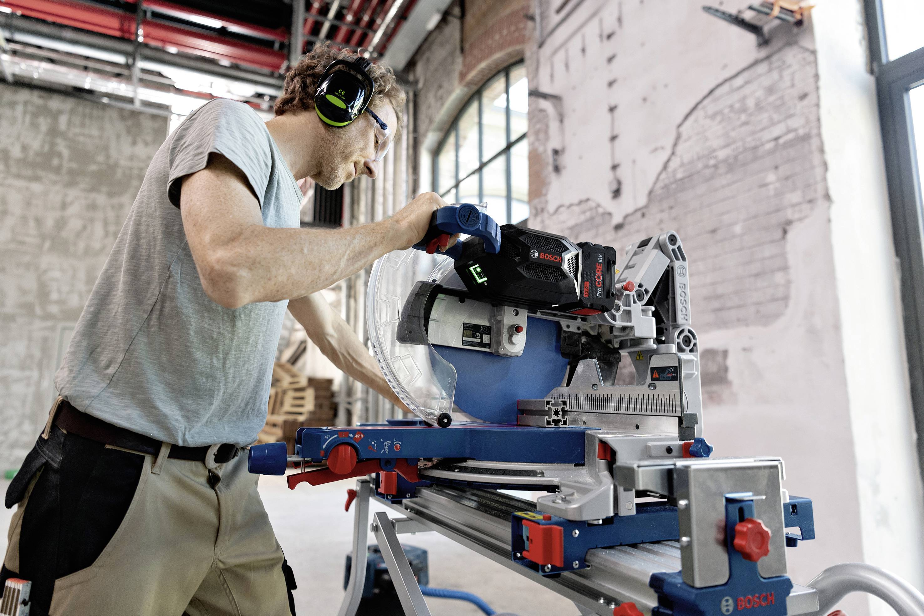 Eine Person arbeitet mit Gehörschutz an einer elektrischen Gehrungssäge in einer industriellen Werkstattumgebung mit sichtbaren Ziegelwänden.