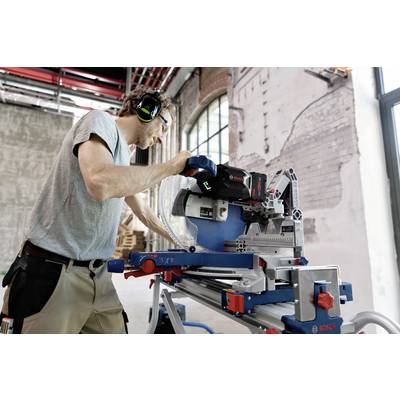 Eine Person arbeitet mit Gehörschutz an einer elektrischen Gehrungssäge in einer industriellen Werkstattumgebung mit sichtbaren Ziegelwänden.
