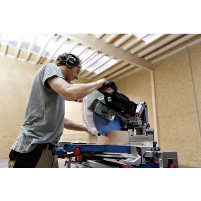 Ein Mann arbeitet in einer Werkstatt mit Kappsäge, schneidet Holz entlang einer markierten Linie. Er trägt Gehörschutz.