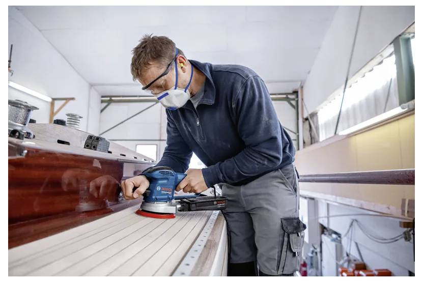 Eine Person mit Schutzausrüstung verwendet einen Exzenterschleifer, um das Holzdeck eines Bootes im Innenraum präzise und detailliert zu glätten.