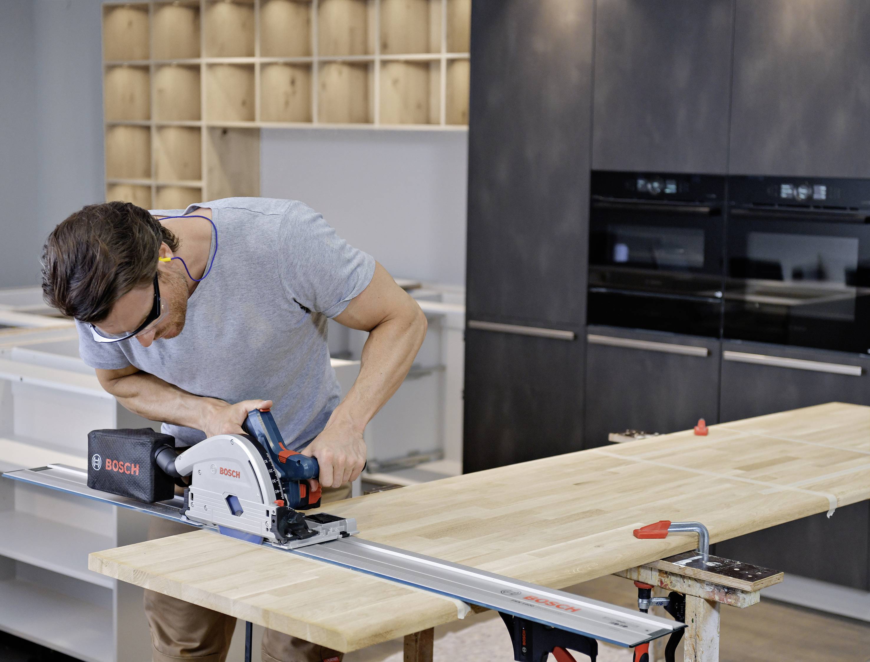 Ein Mann sägt ein Holzbrett in einer Werkstatt mit einer Handkreissäge. Im Hintergrund sind Regale und Küchengeräte.