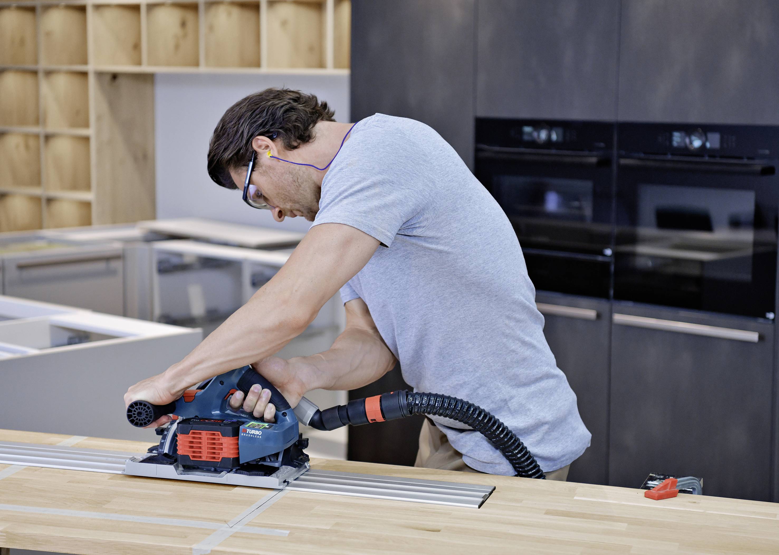 Ein Mann arbeitet in einer Werkstatt an einer Arbeitsplatte mit einer elektrischen Säge. Im Hintergrund sind Schränke zu sehen.