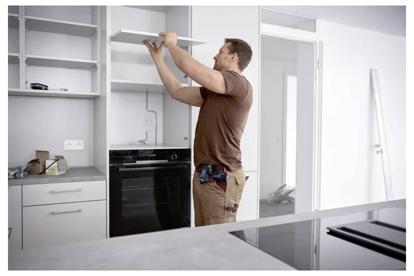 Ein Mann installiert ein Wandschrankregal in einer modernen Küche, während er mit einem Elektrowerkzeug nach oben greift und neben eingebauten Geräten steht.
