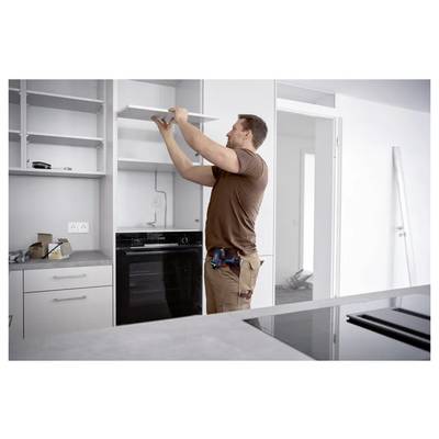 Ein Mann installiert ein Wandschrankregal in einer modernen Küche, während er mit einem Elektrowerkzeug nach oben greift und neben eingebauten Geräten steht.