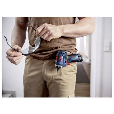 Arbeiter in beigefarbener Hose mit einem schnurlosen Bohrer an der Hüfte befestigt, der eine Schutzbrille in einer Hand hält.