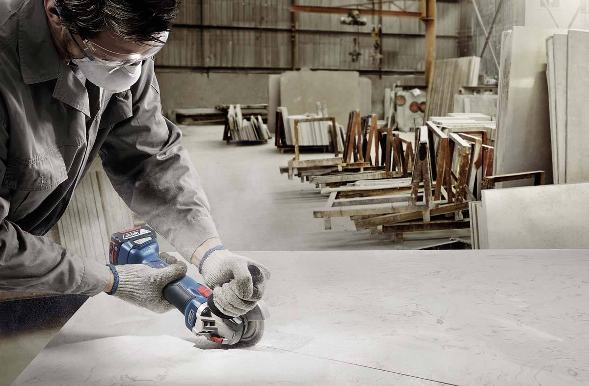 Eine Person in Schutzkleidung schneidet mit einem elektrischen Werkzeug eine Marmorplatte in einer Fabrikhalle. Holzregale im Hintergrund.