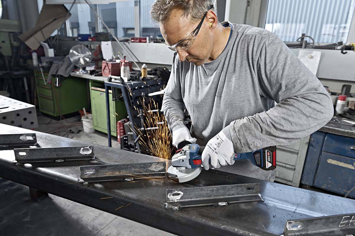 Ein Mann mit Schutzbrille schleift Metall in einer Werkstatt. Funken fliegen von der Schleifscheibe. Um ihn herum stehen Werkzeuge und Geräte.