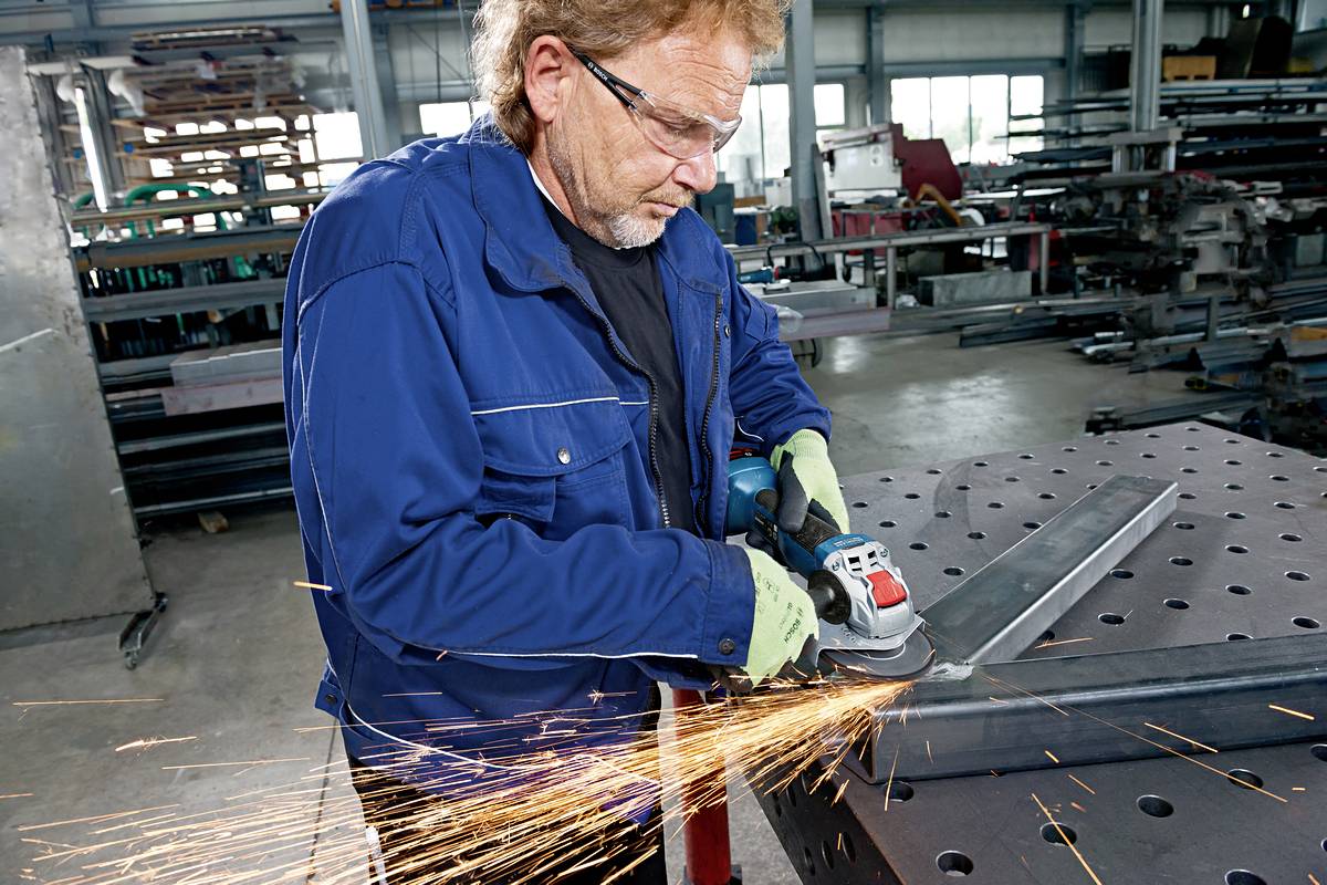 Ein Mann schleift Metall in einer Werkstatt; Funken fliegen. Er trägt Schutzbrille und Handschuhe; Sicherheitsmaßnahmen sind vorhanden.