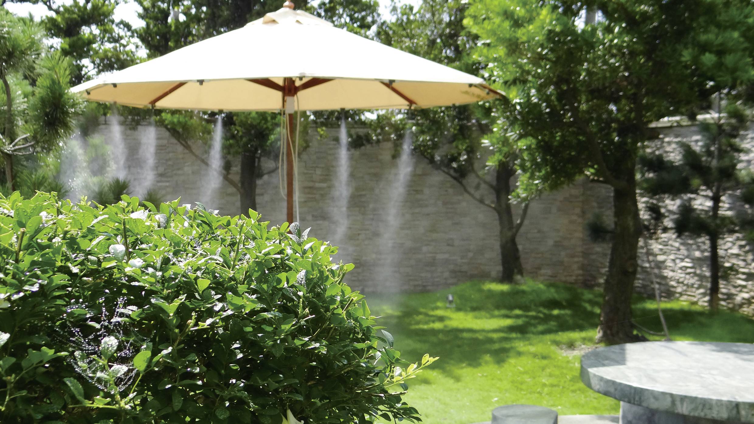 Ein großer Sonnenschirm spendet Schatten in einem grünen Garten mit Bäumen und einem runden Tisch daneben.