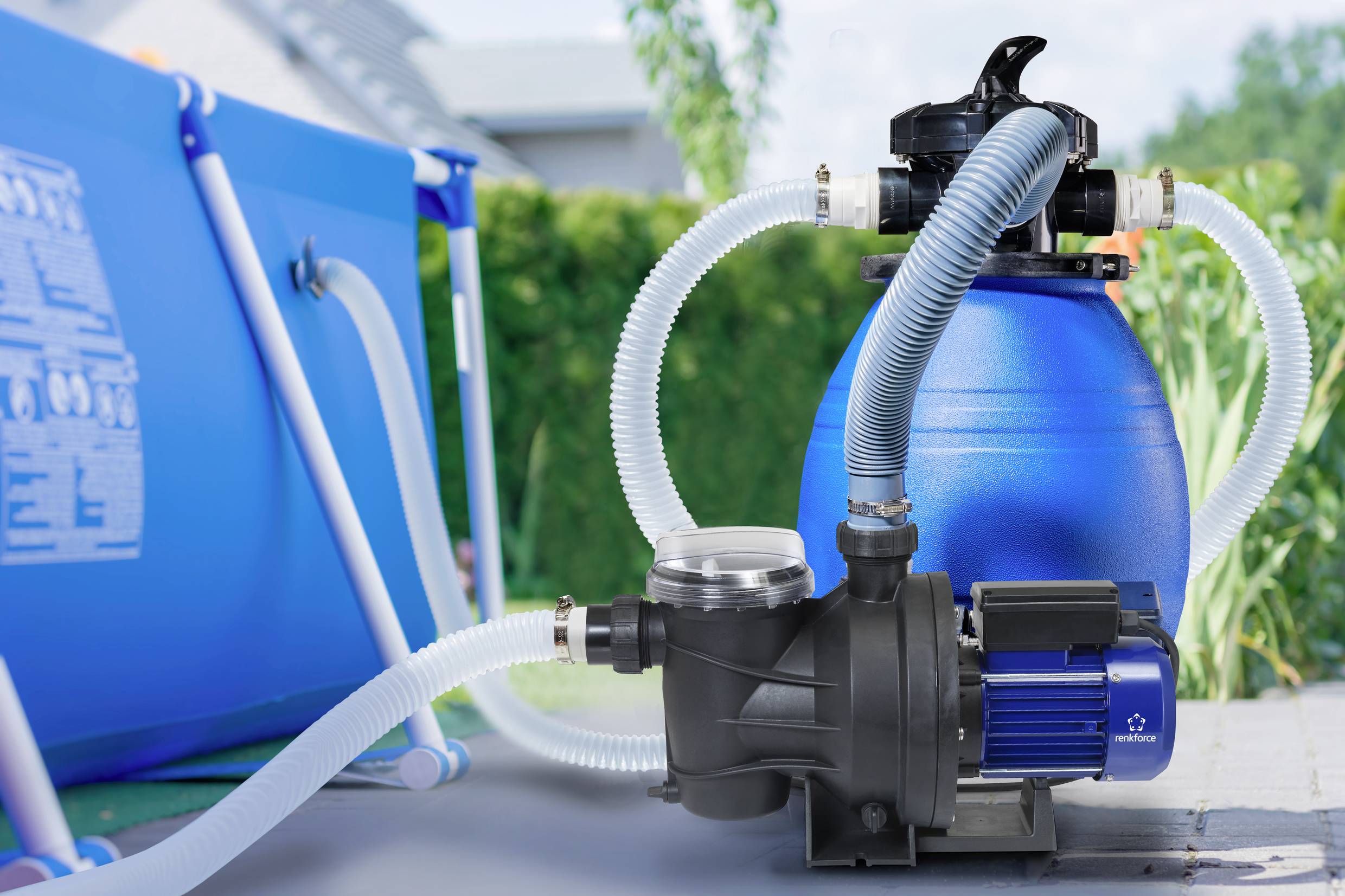Blaues Sandfilteranlage-Set neben einem Pool, verbunden mit Schläuchen, vor einem Gartenhintergrund.