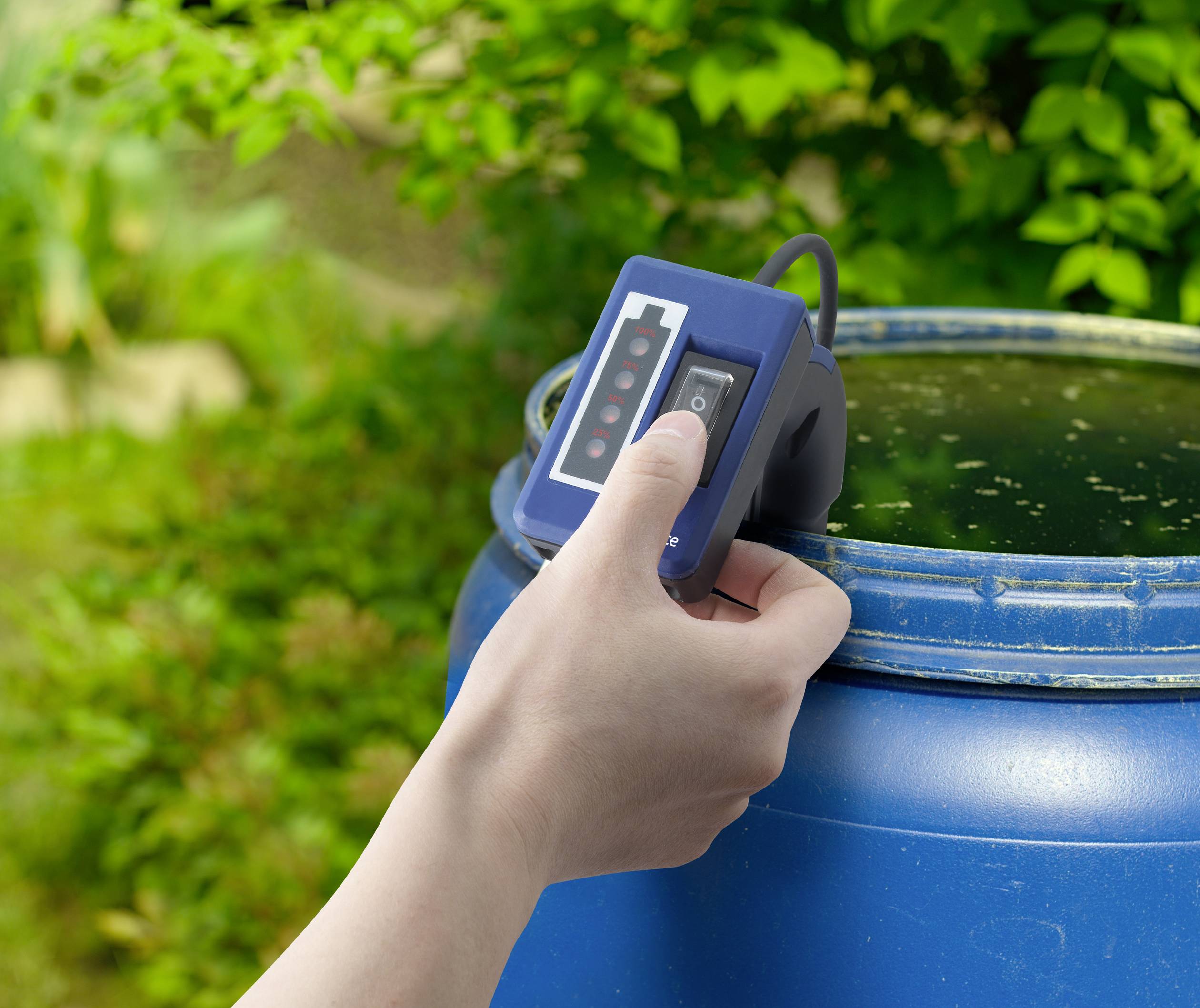 Eine Hand hält ein digitales Messgerät über ein blaues Fass im Freien, um den Wasserstand oder -qualität zu überprüfen. Im Hintergrund ist grüne Vegetation zu sehen.