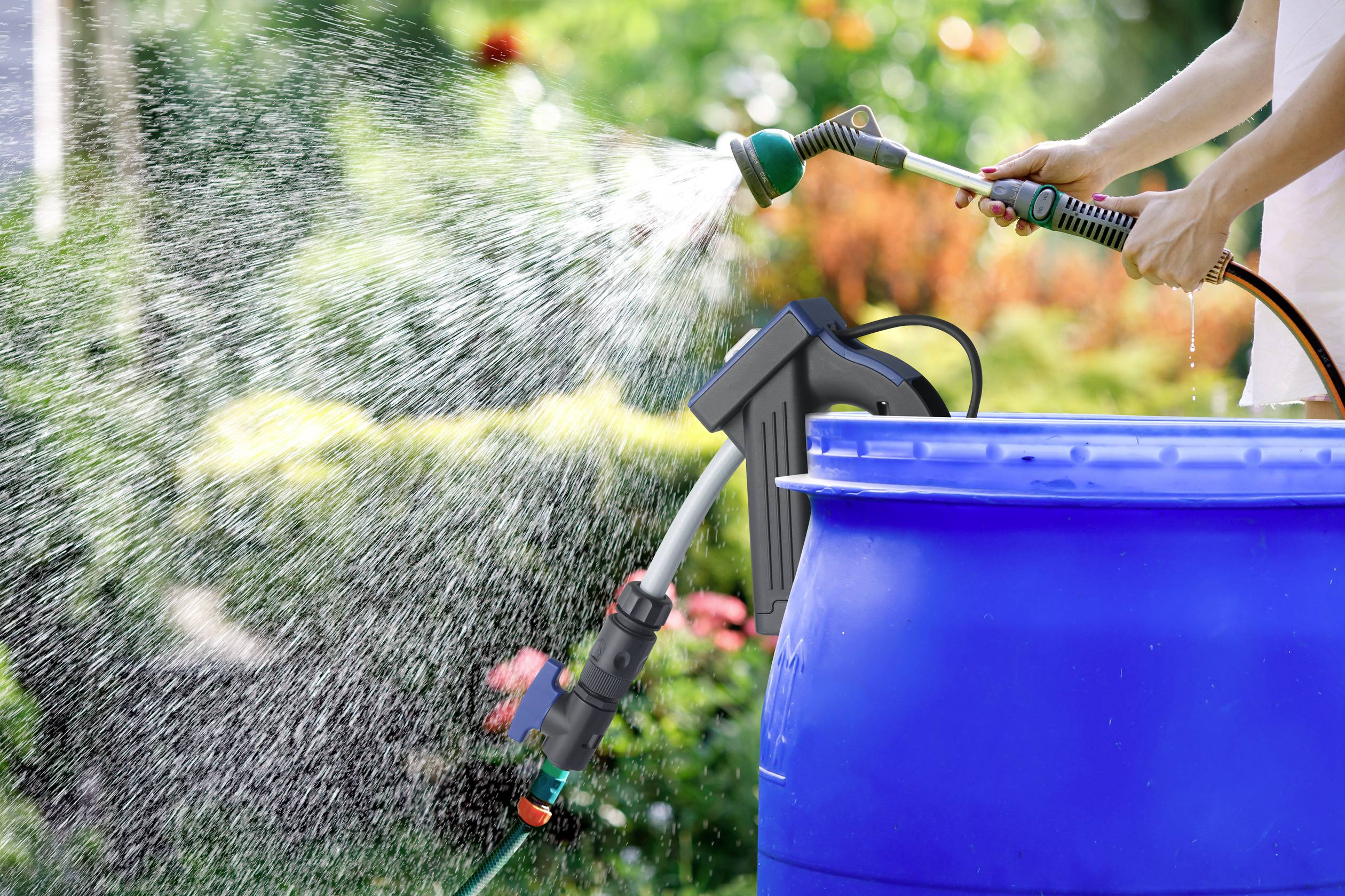 Person gießt mit Gartenschlauch Wasser in eine blaue Regentonne. Im Hintergrund ist ein grüner Garten sichtbar.