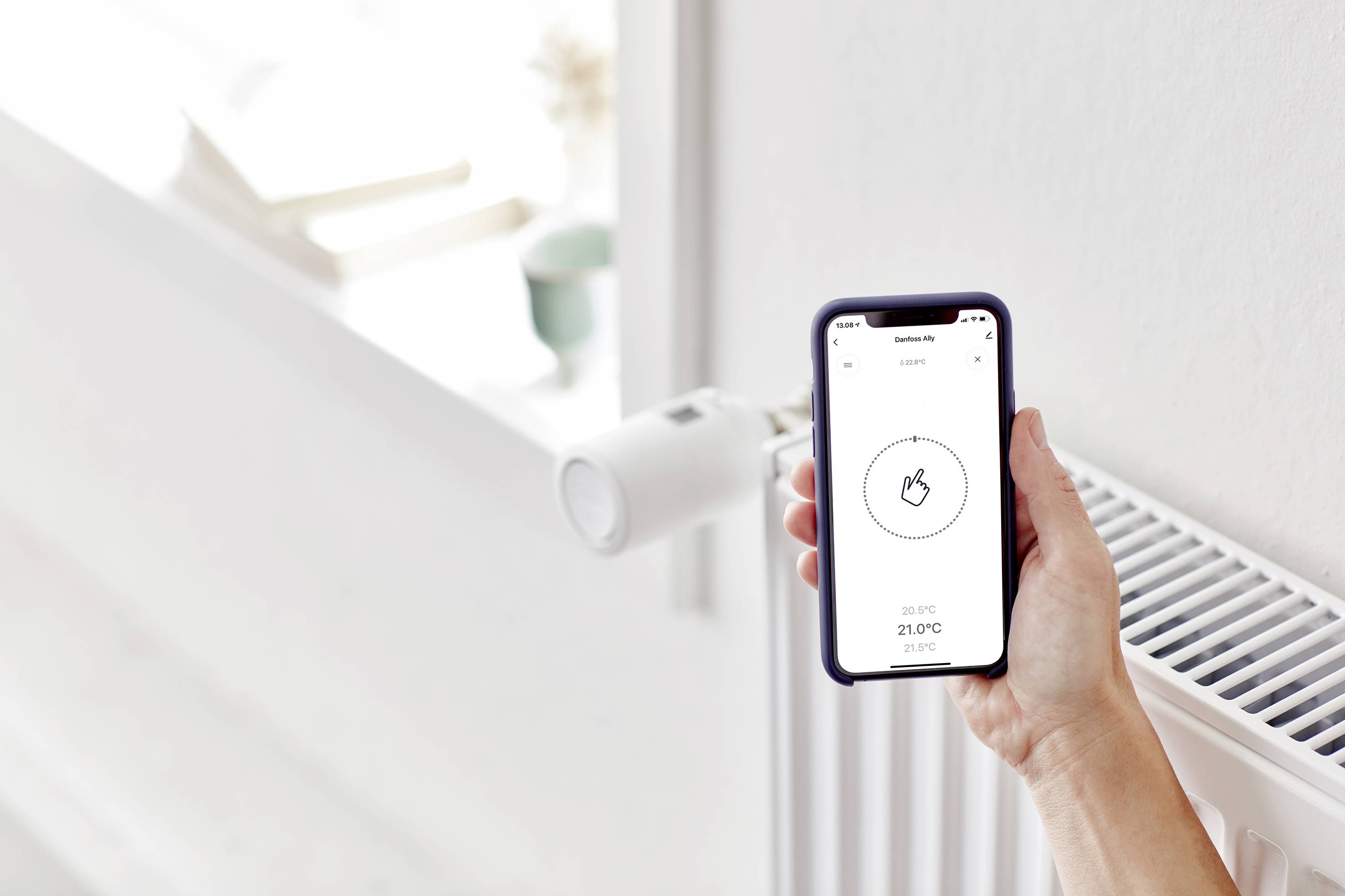 Eine Hand hält ein Smartphone mit einer App zur Heizungssteuerung. Im Hintergrund ist ein Heizkörper mit einem smarten Thermostat zu sehen.