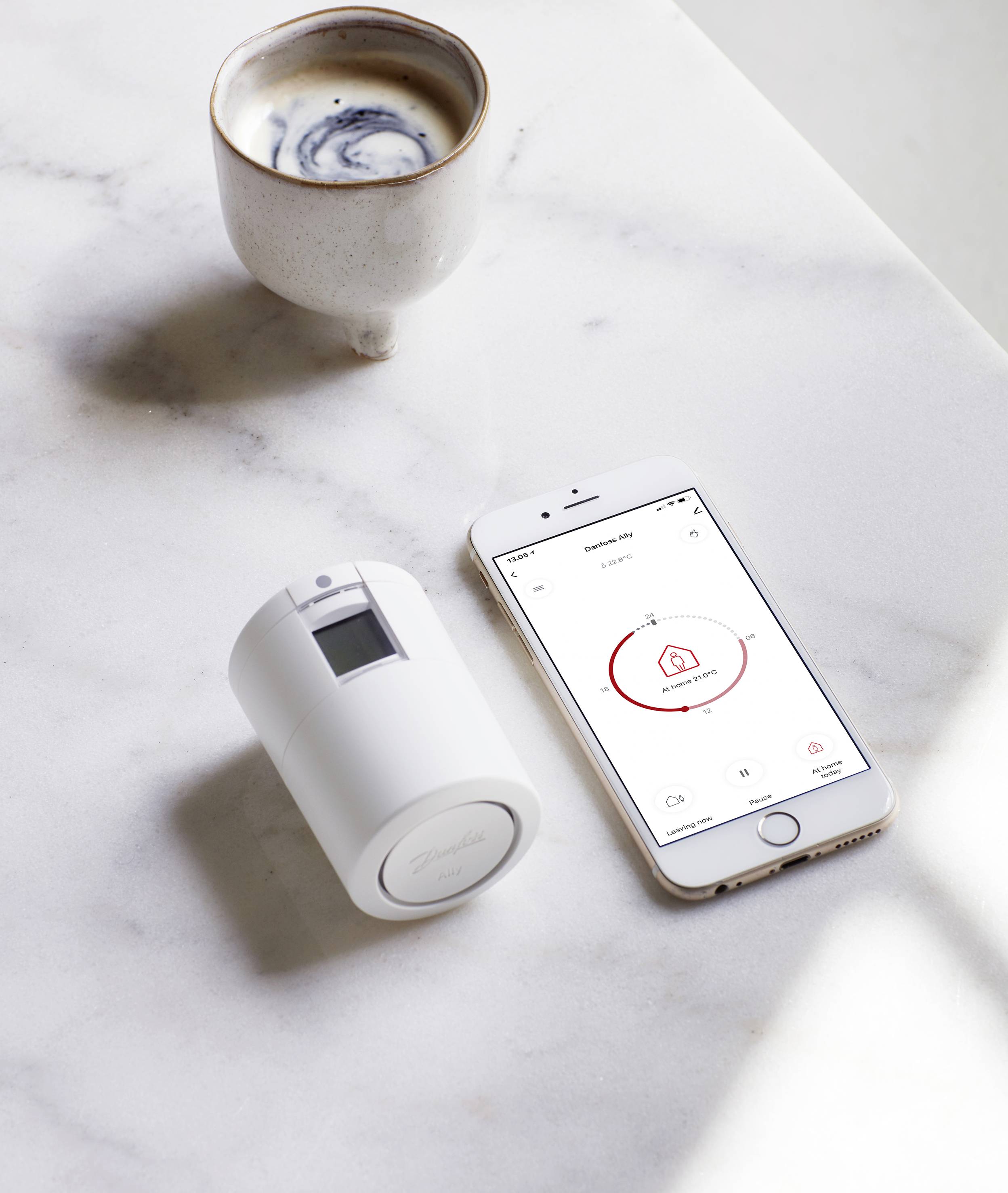 Ein weißer Becher Kaffee, ein Smartphone mit einer geöffneten App und ein weißes, zylindrisches Gerät liegen auf einem Marmortisch.