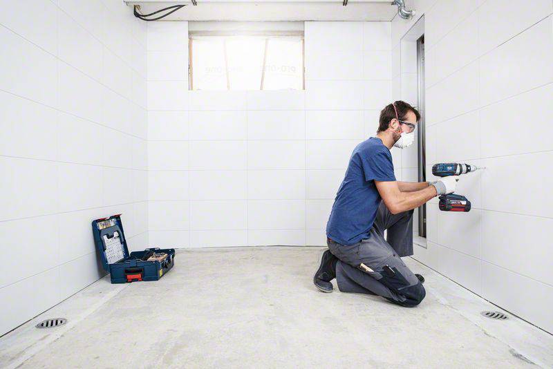 Eine Person kniet in einem leeren Raum und arbeitet mit einer Bohrmaschine an der Wand, Werkzeugkasten links auf dem Boden.