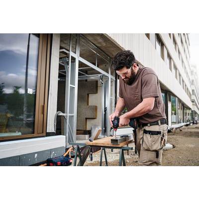 Ein Handwerker bohrt ein Loch in ein Metallstück auf einem Arbeitstisch vor einem modernen Gebäude. Er trägt Arbeitskleidung.