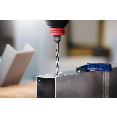 Eine Bohrmaschine bohrt mit einem Metallbohrer in ein rechteckiges Aluminiumrohr. Späne fallen um die Bohrstelle herum.
