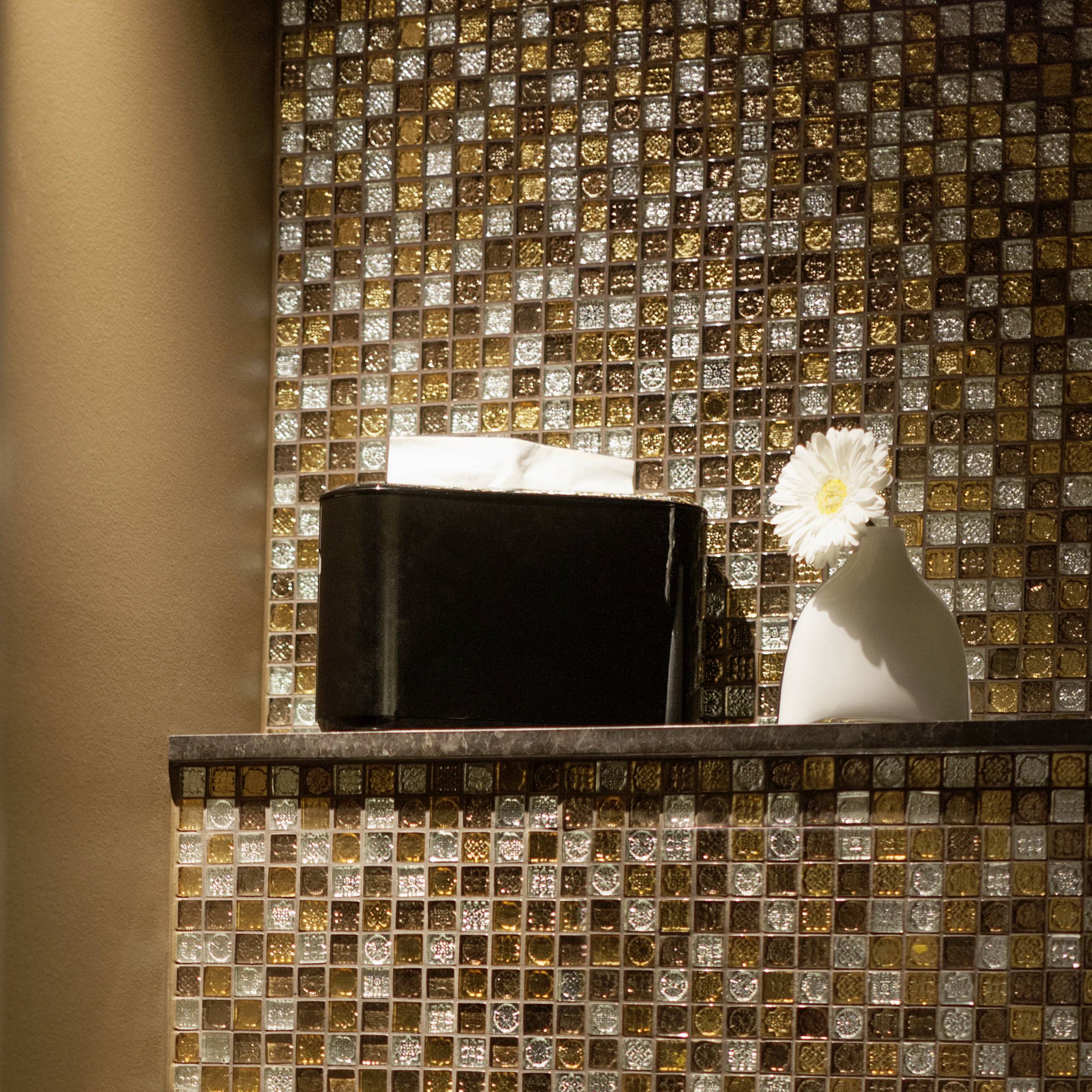 Ein Regal mit einer schwarzen Box und einer weißen Vase mit einer Blume, beleuchtet vor einer glitzernden Mosaikwand.