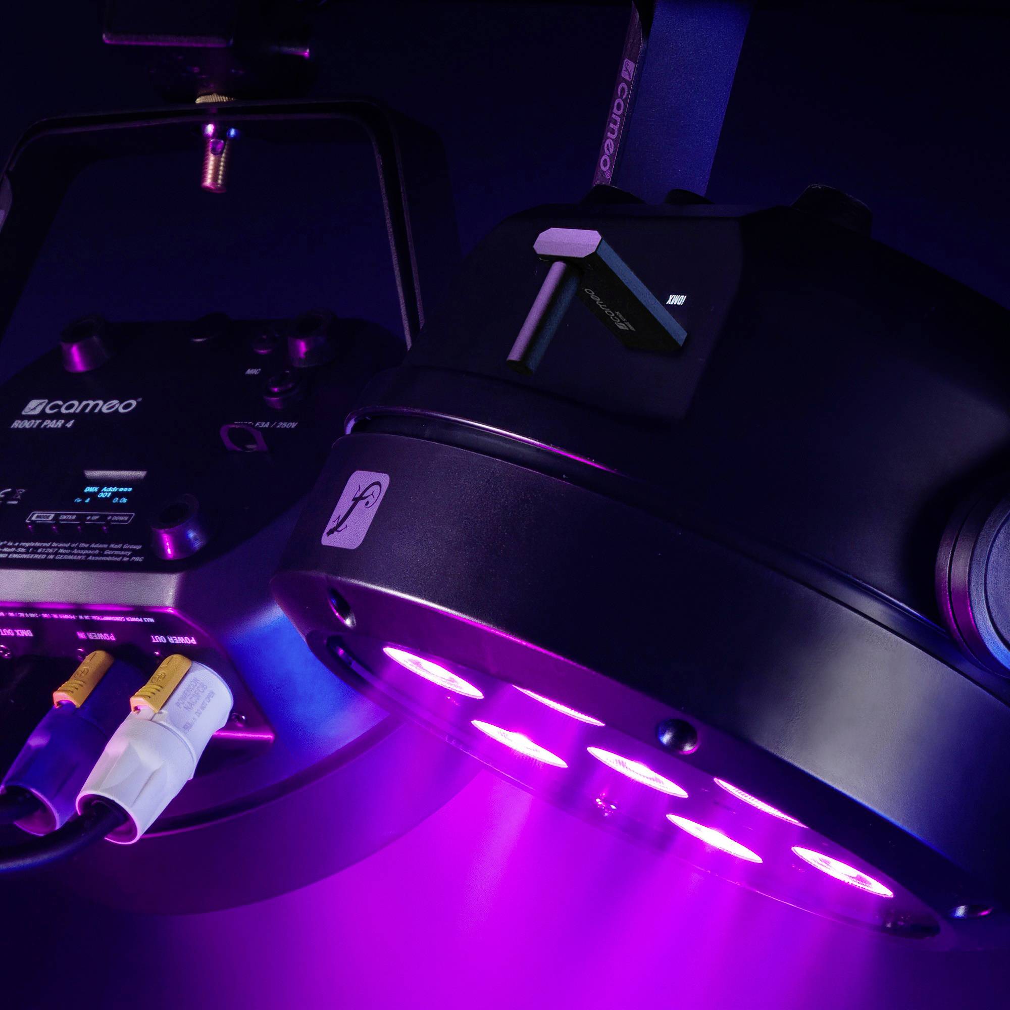 Ein schwarzer, runder LED-Scheinwerfer, der violett leuchtet, hängt von einer metallischen Halterung. Im Hintergrund sind Kabel sichtbar.