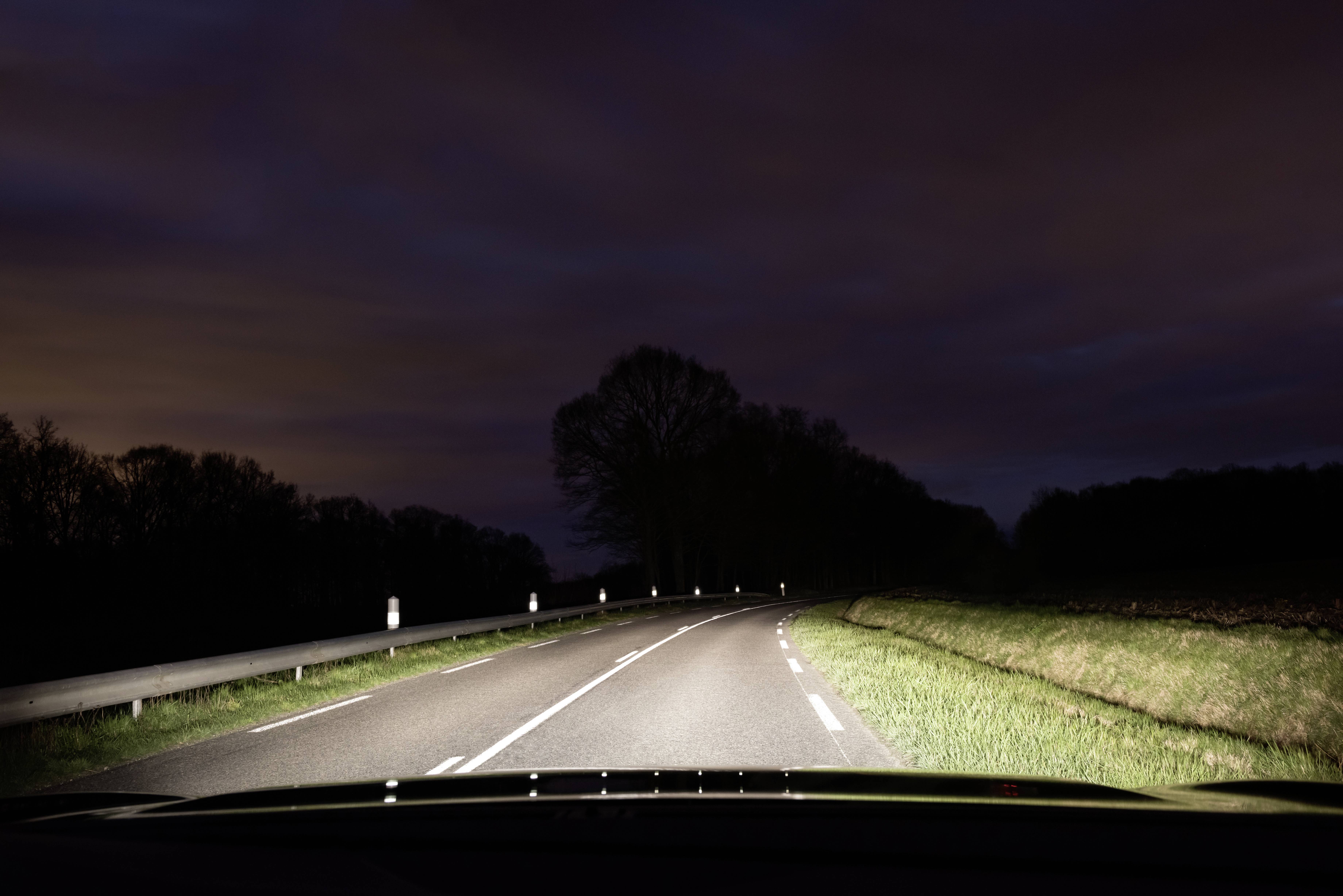 Straße bei Nacht beleuchtet von Autoscheinwerfern, führt in eine Kurve mit Bäumen und Wiesen an den Seiten. Dunkler, wolkiger Himmel.